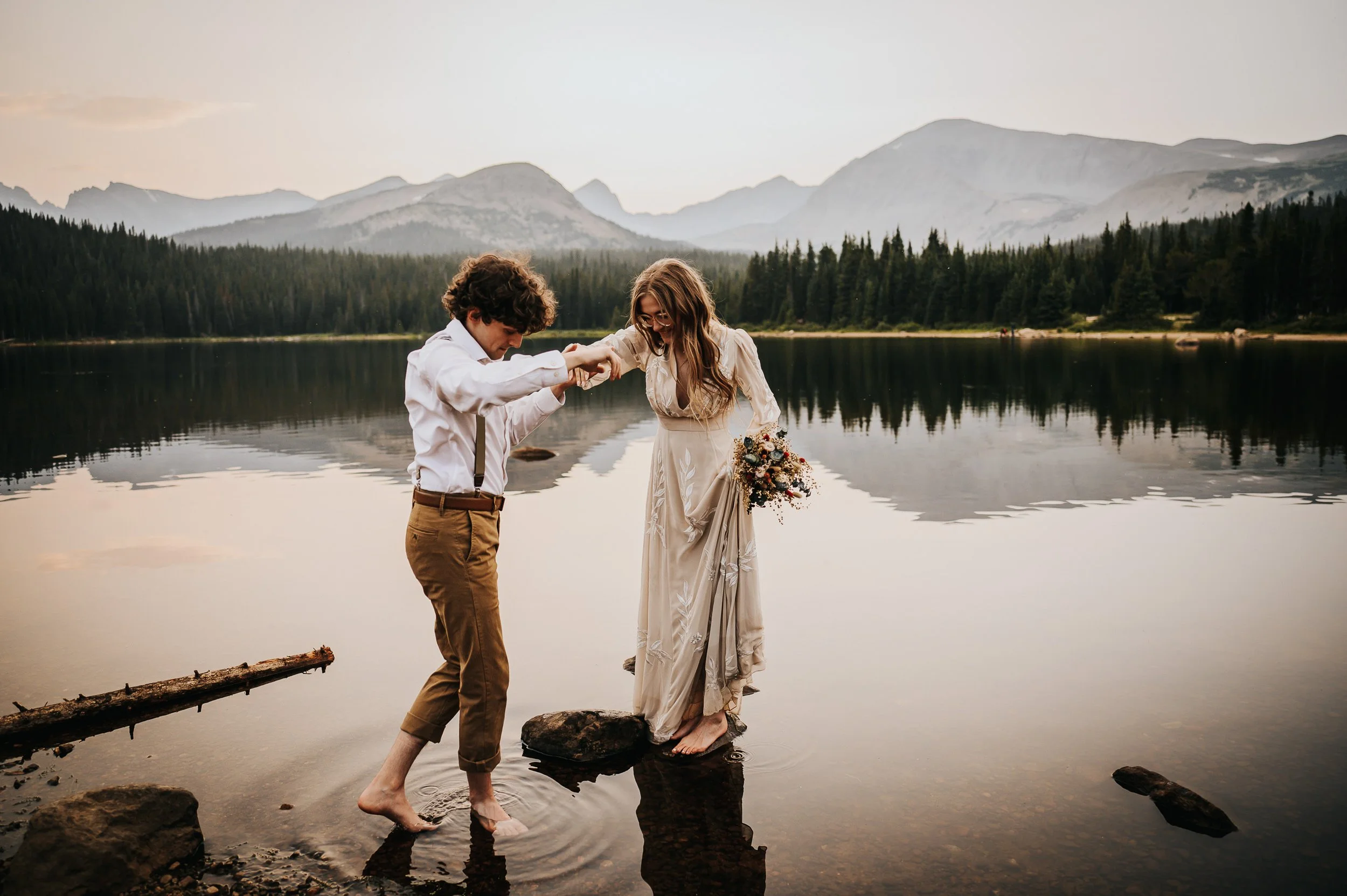 A groom helps his bride with something at the edge of a still mountain lake, golden hour light.