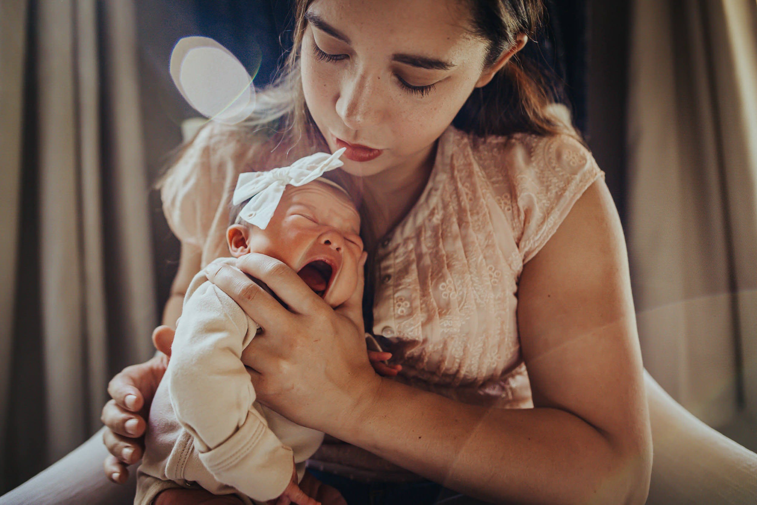 A mother gazes down at her newborn with quiet tenderness in soft window light.