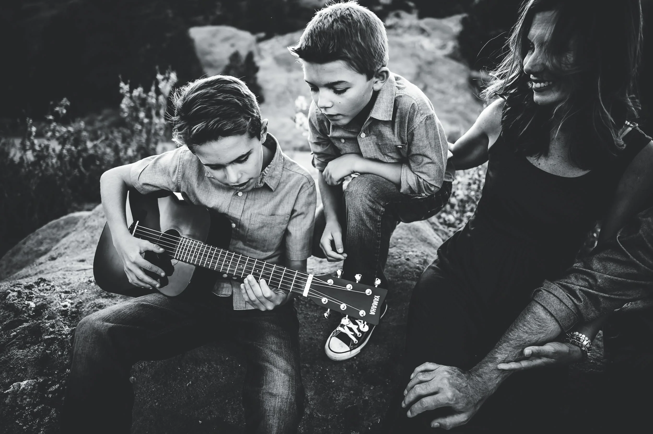 A son plays acoustic guitar on rocks while brother and mom sit nearby, black and white portrait.