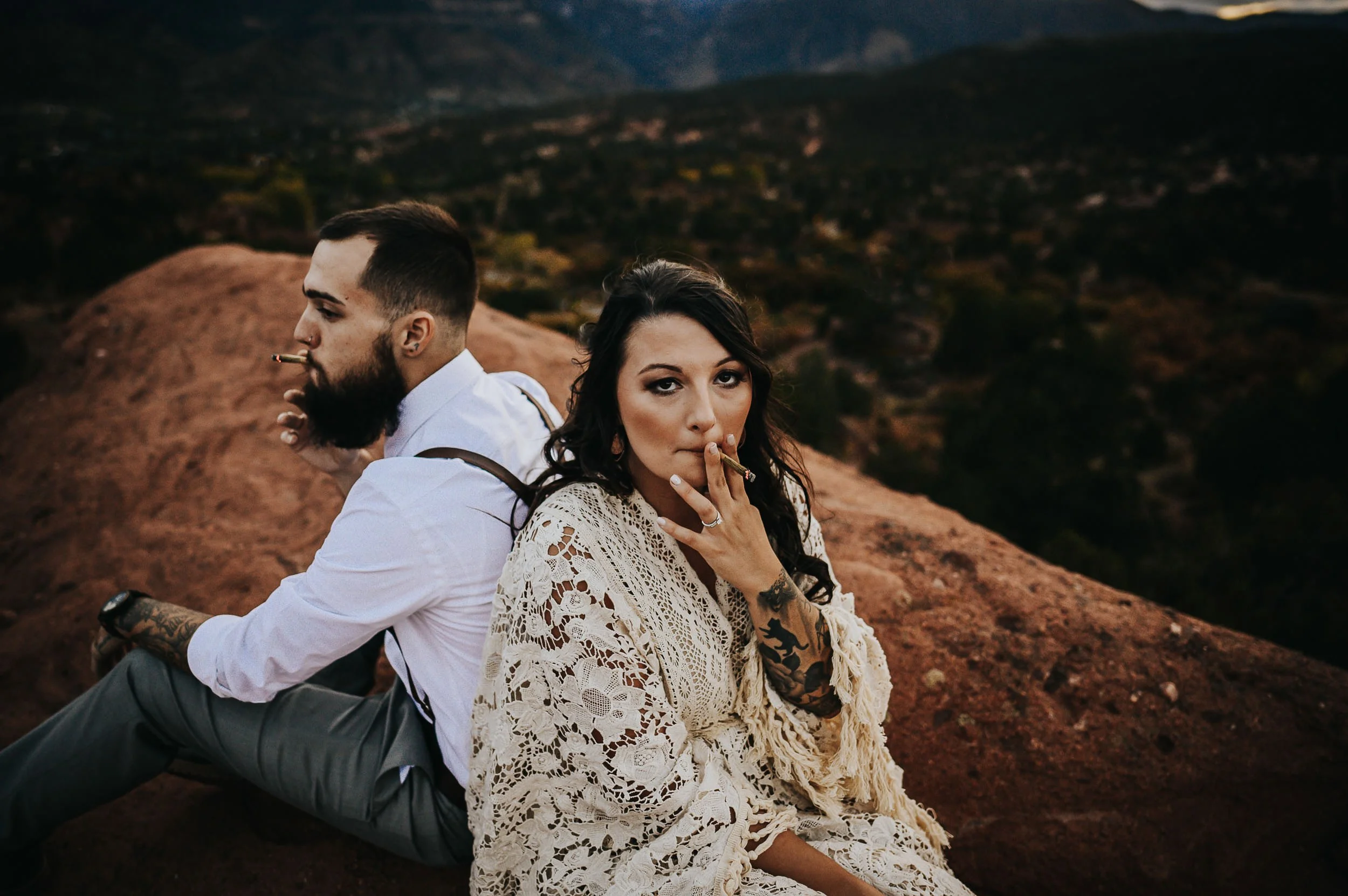 A couple sits close on red rocks, she in a boho lace dress, he in white with suspenders, faces close.