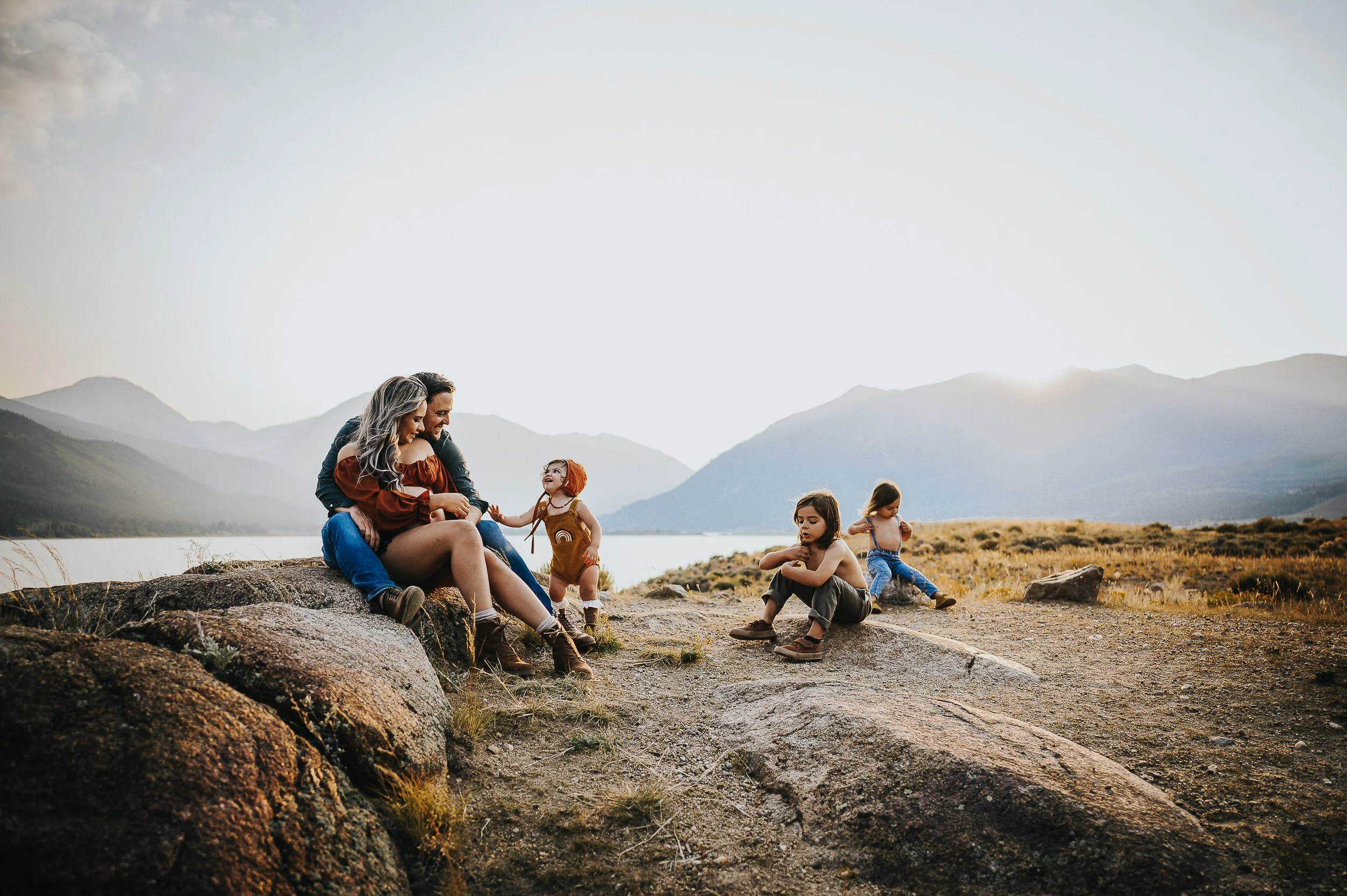 Family of five sitting on rocks by a Colorado mountain lake, coordinated in rust, mustard, and navy tones with mountains in the background.