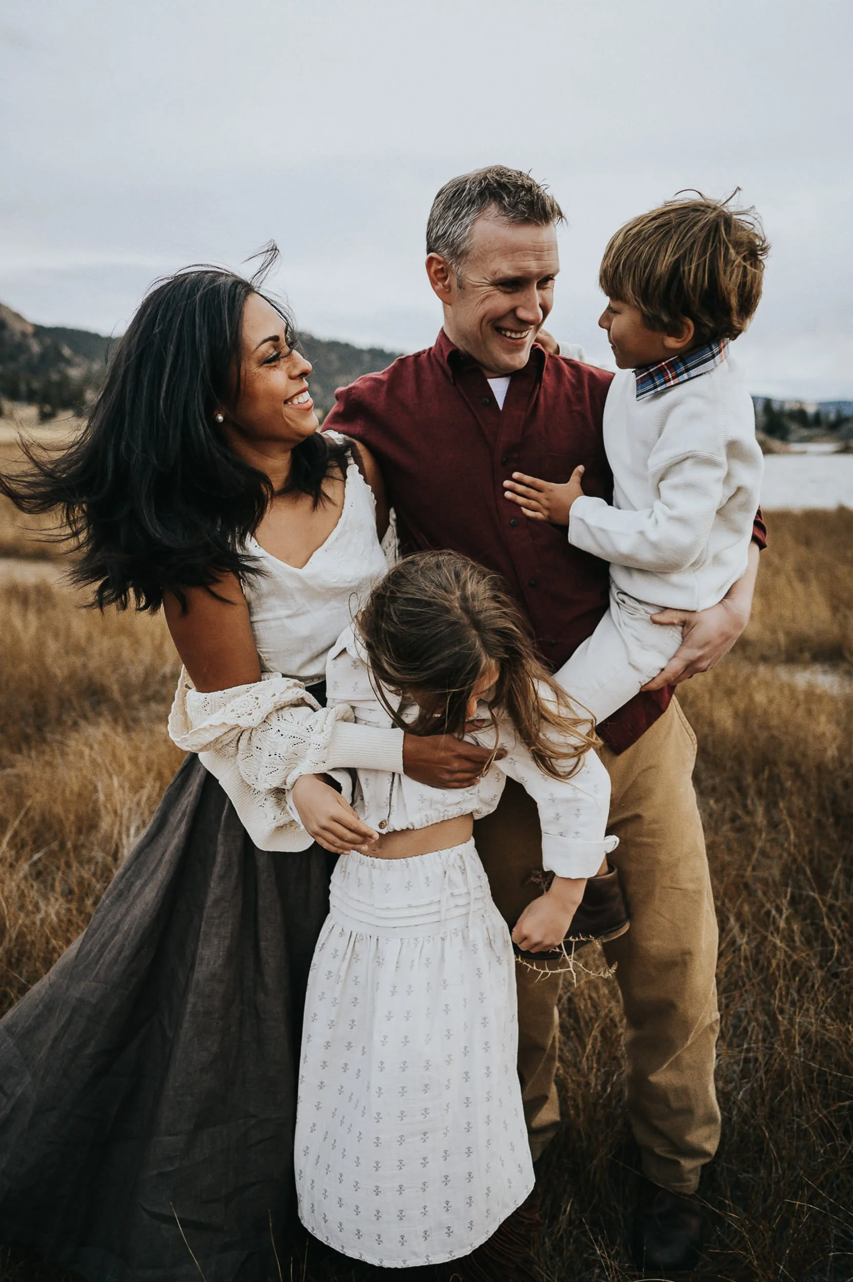 Family of four with dad in olive sweater and mom in cream, dad playfully lifting child while older child watches.