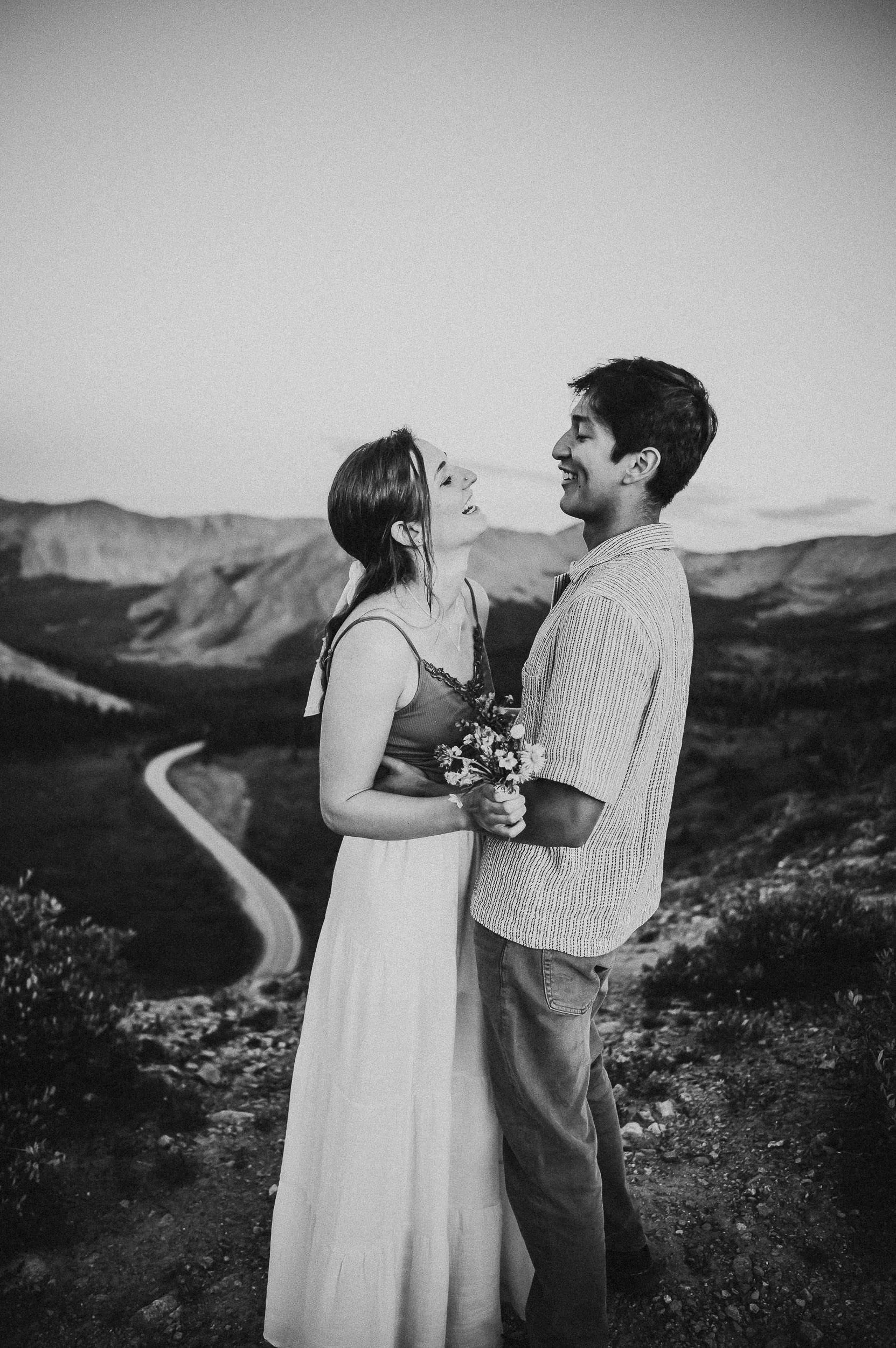 Couple holding wildflowers with a winding mountain road stretching behind them, black and white.