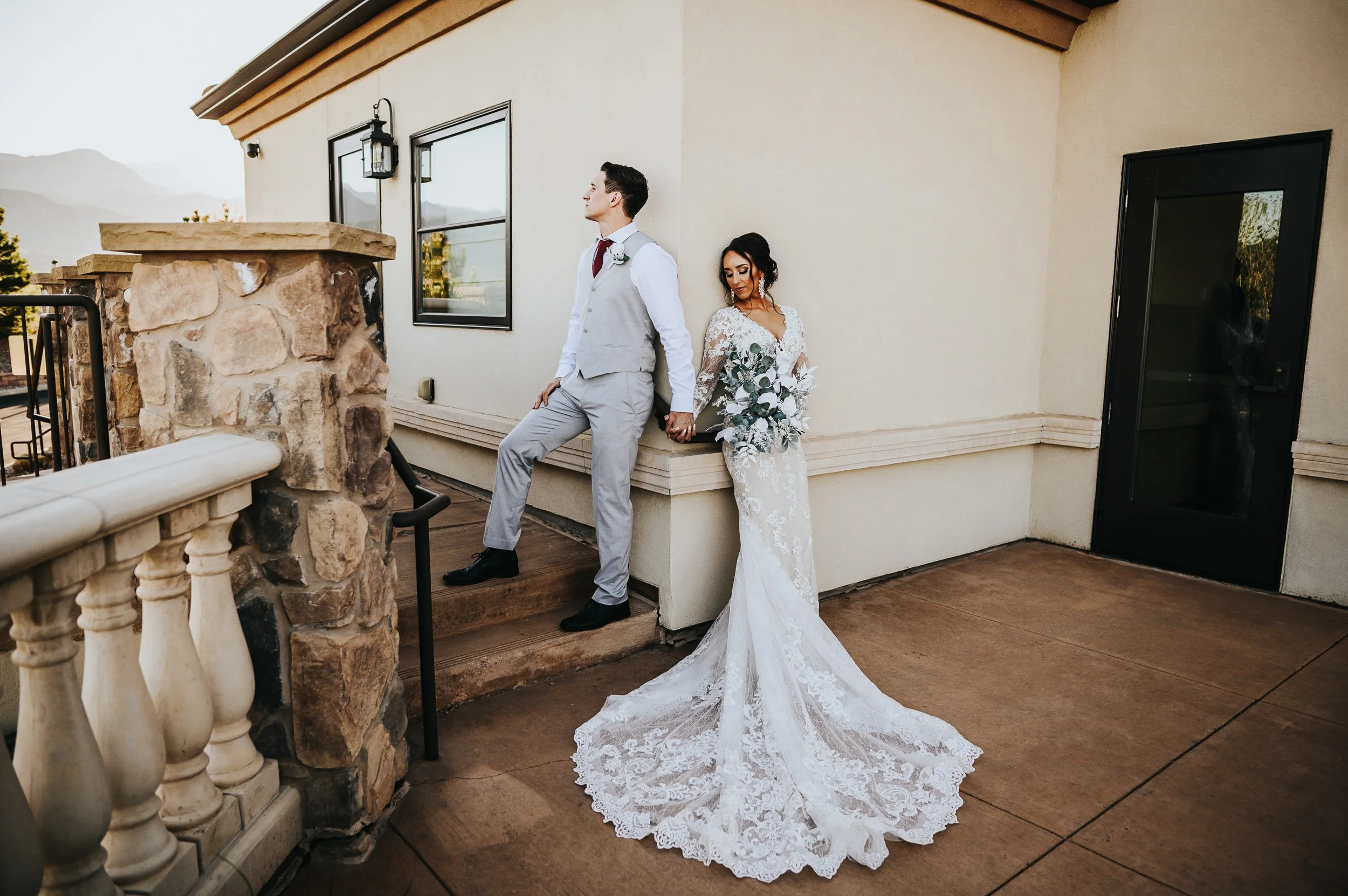 Bride and groom posing on stone steps outside a wedding venue, bride in a fitted lace gown with a long train.