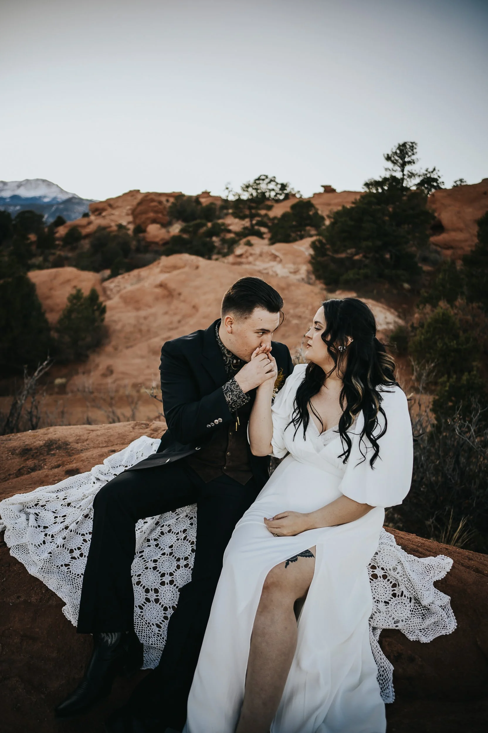 A bride and groom sit together on a hillside in the Colorado mountains, he in a suit, she in a full lace gown.