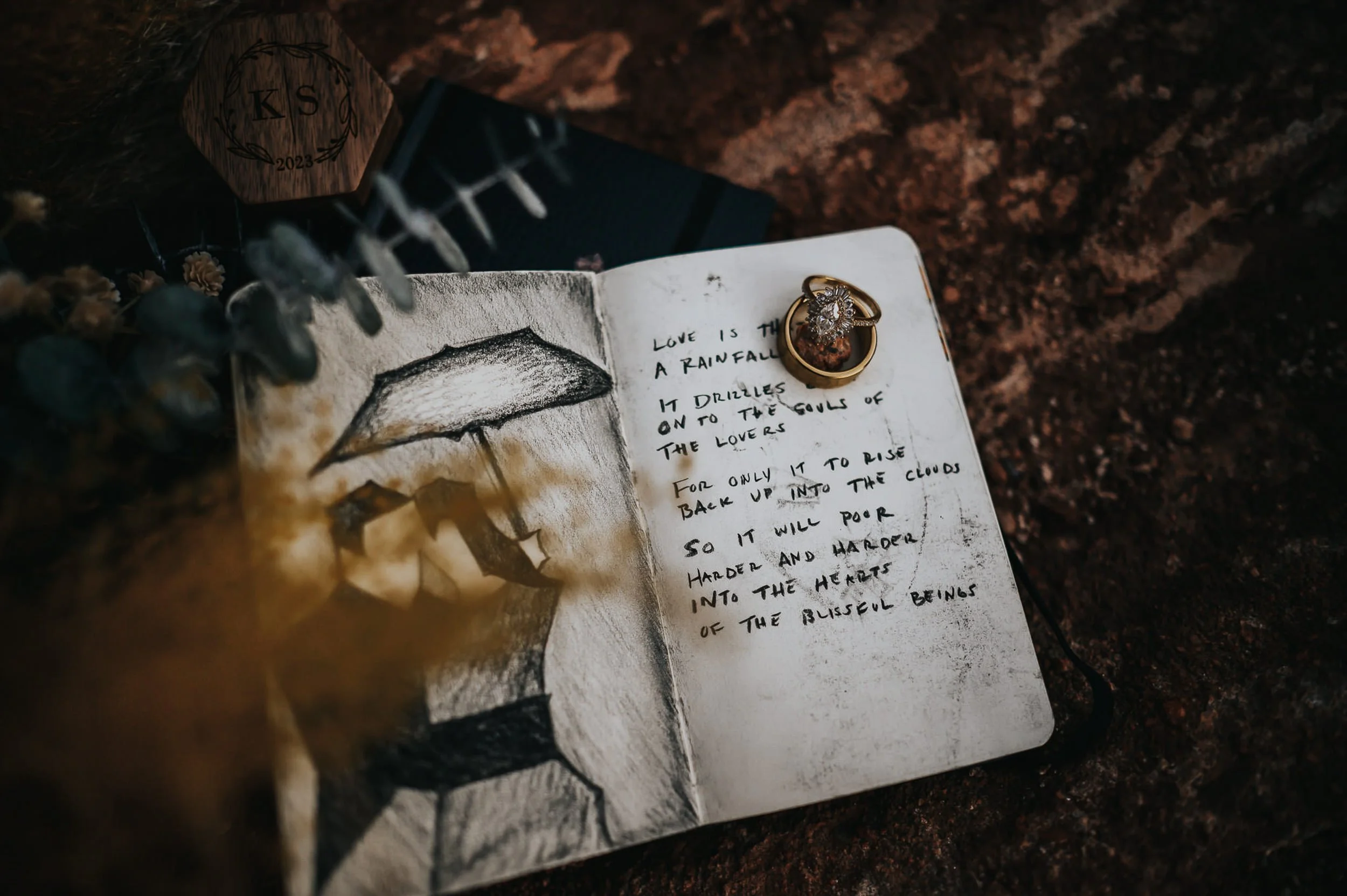 A handwritten vow book lies open on a weathered surface, dried flowers alongside it.