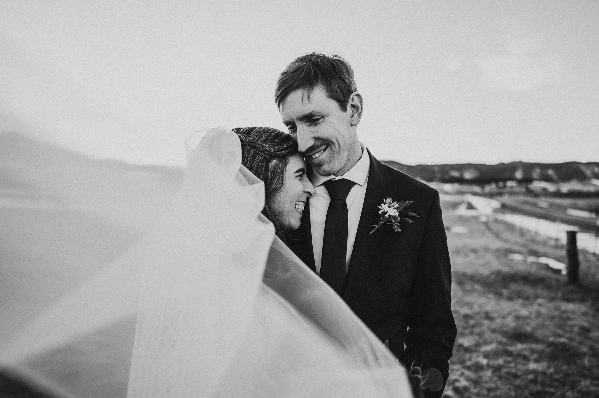A groom in a suit smiles down tenderly at his bride, black and white closeup portrait.