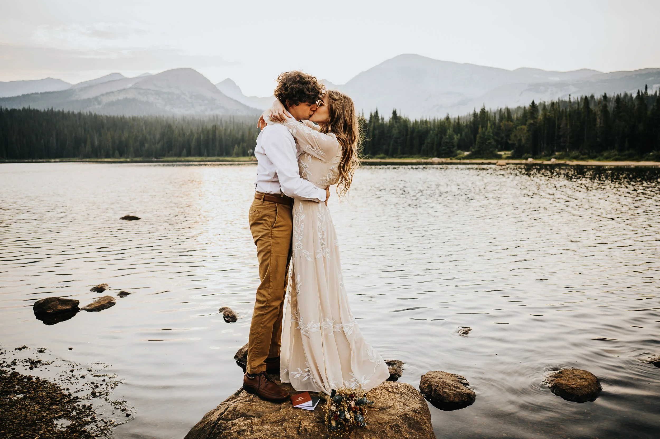 Couple sharing a kiss on rocks beside a calm mountain lake with forested hills behind them.