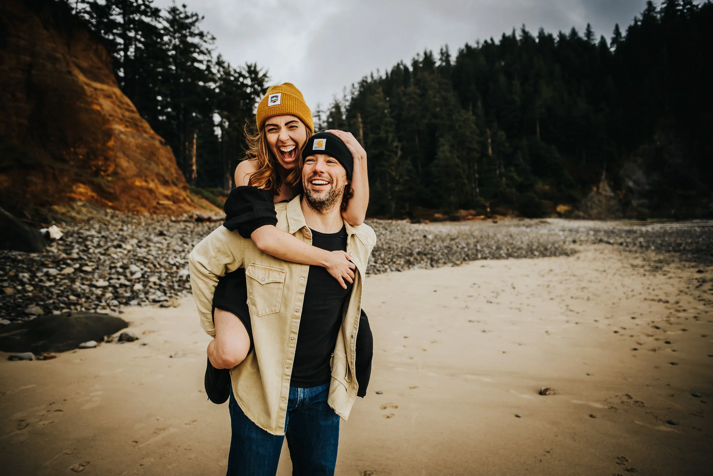 A couple laughs together outdoors, he lifts her from behind, both in cozy fall layers.