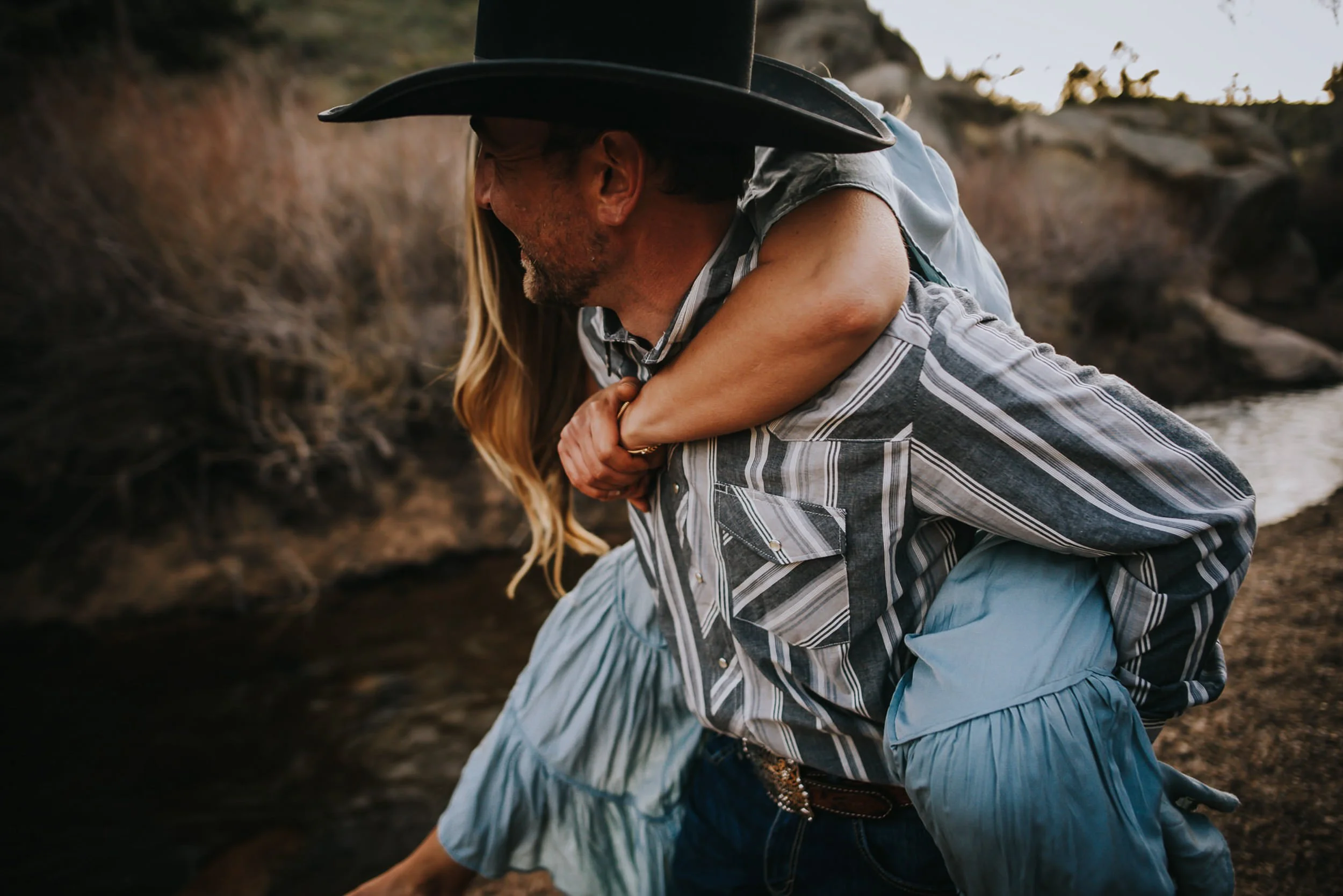A couple in Western attire embrace closely, she wears a blue fringe skirt and cowboy hat.