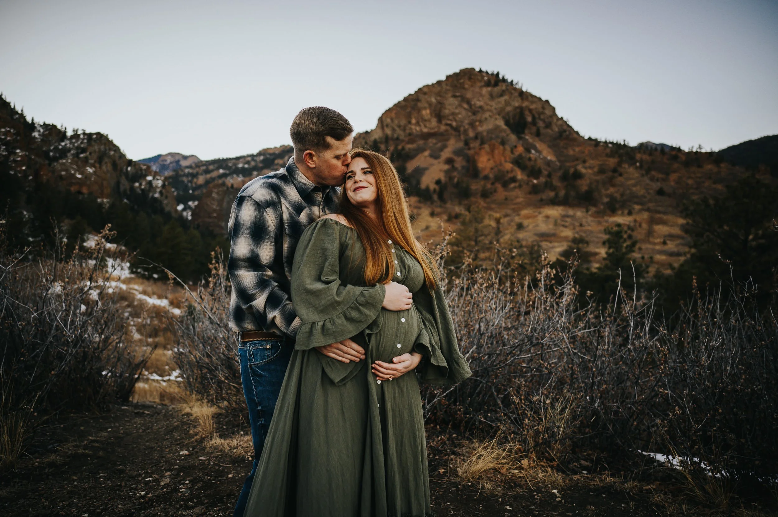 Partner kissing pregnant woman's temple in a golden autumn field with mountains in the distance.