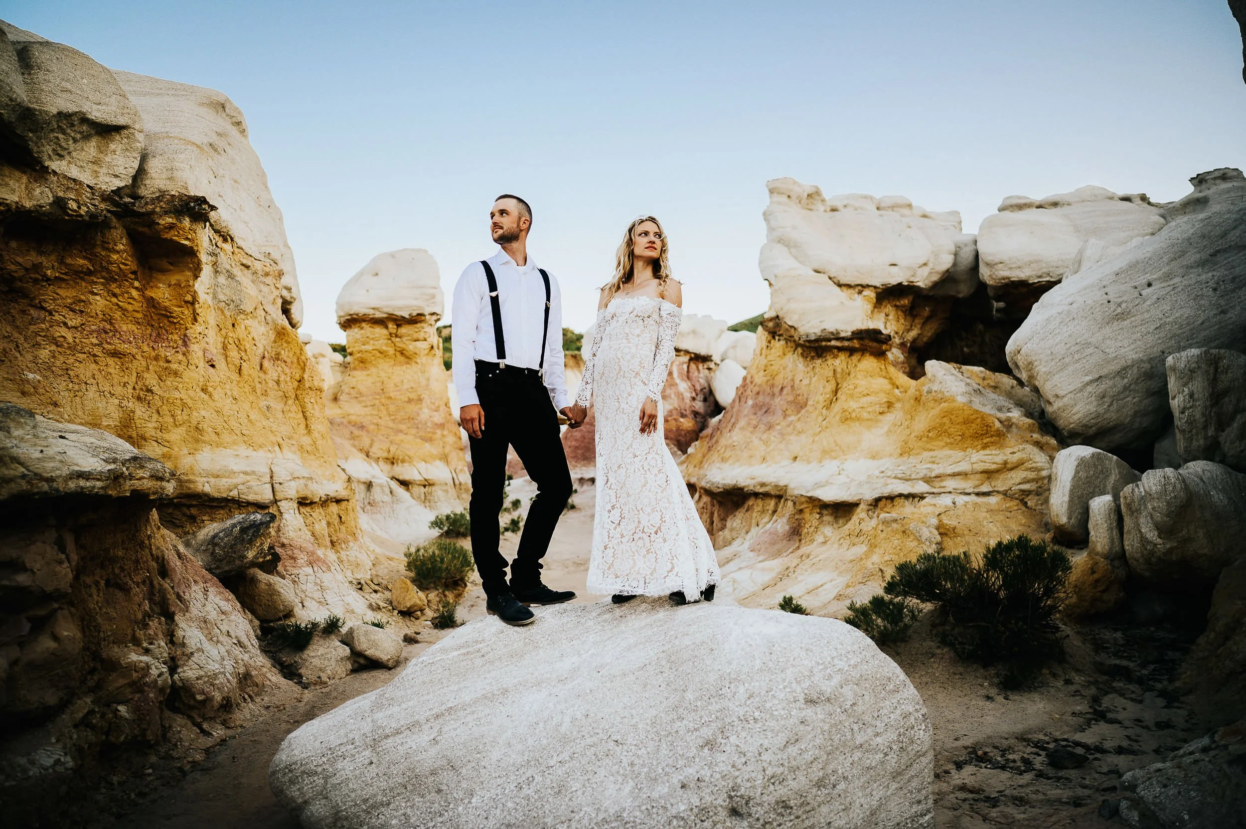 A bride and groom stand together in a white rock landscape, he in suspenders, wide environmental shot.