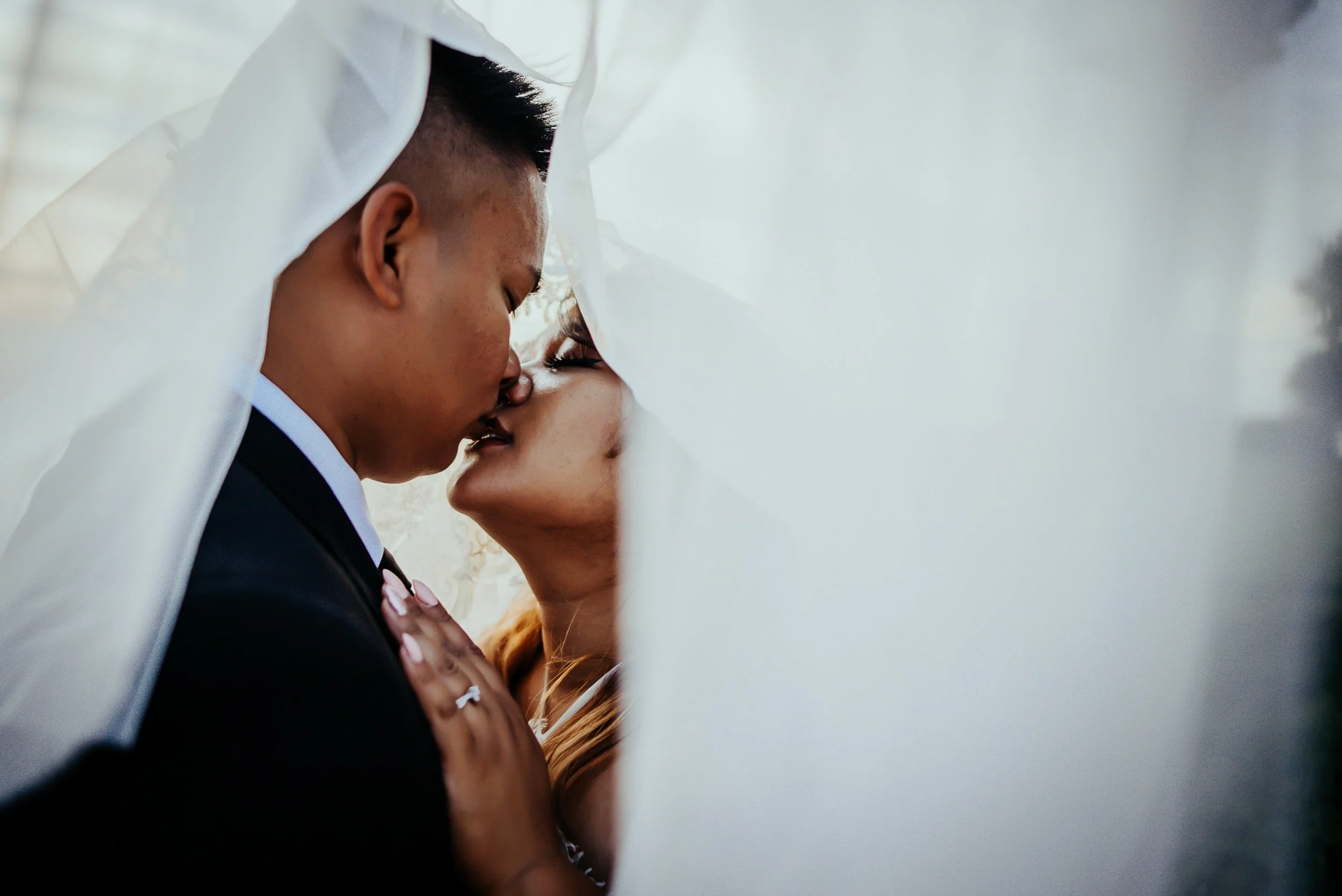 A bride and groom share a nearly-kissing moment, veil floating between their faces, soft light.