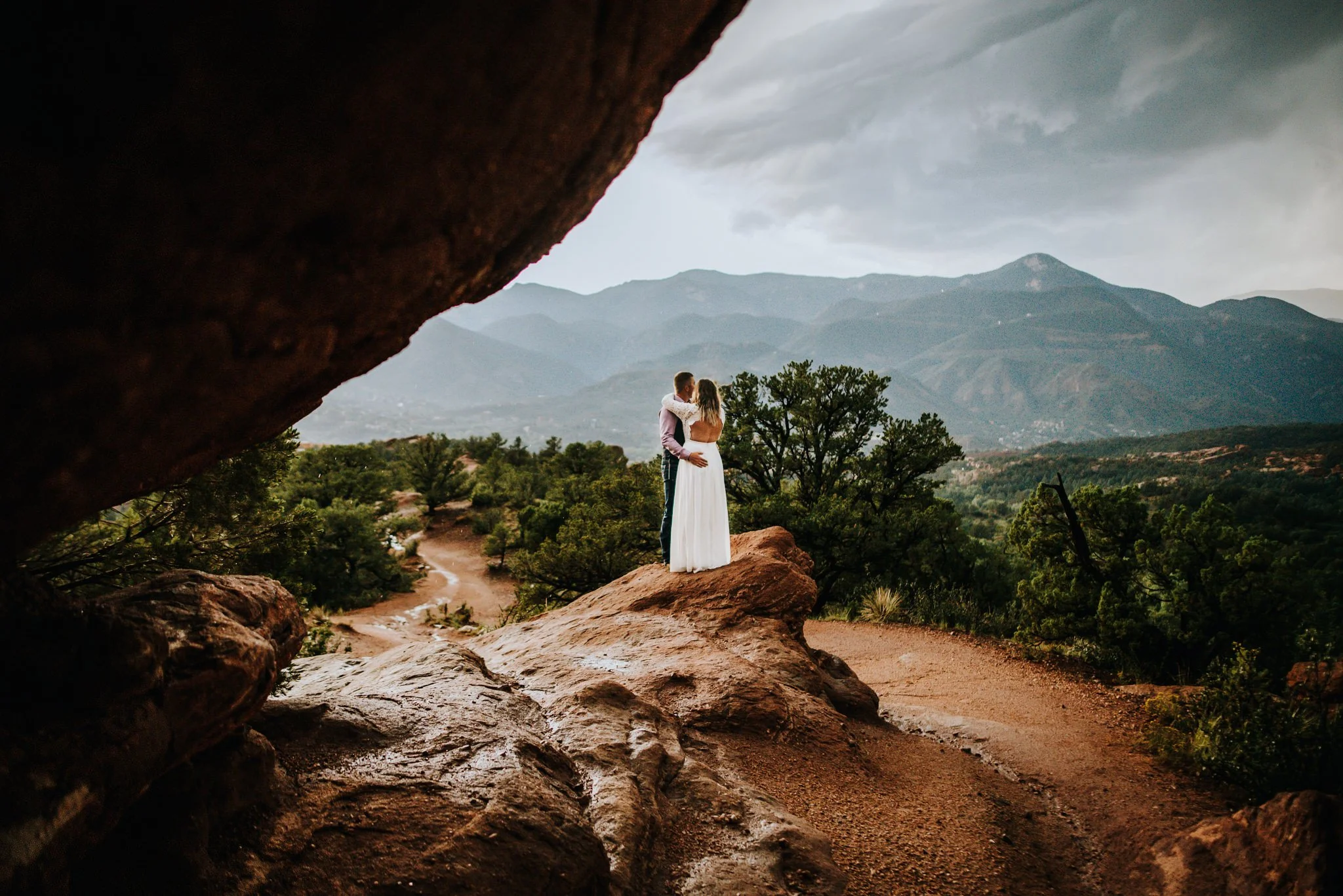 Couple standing together framed by a red rock arch at Garden of the Gods with mountains in the distance.