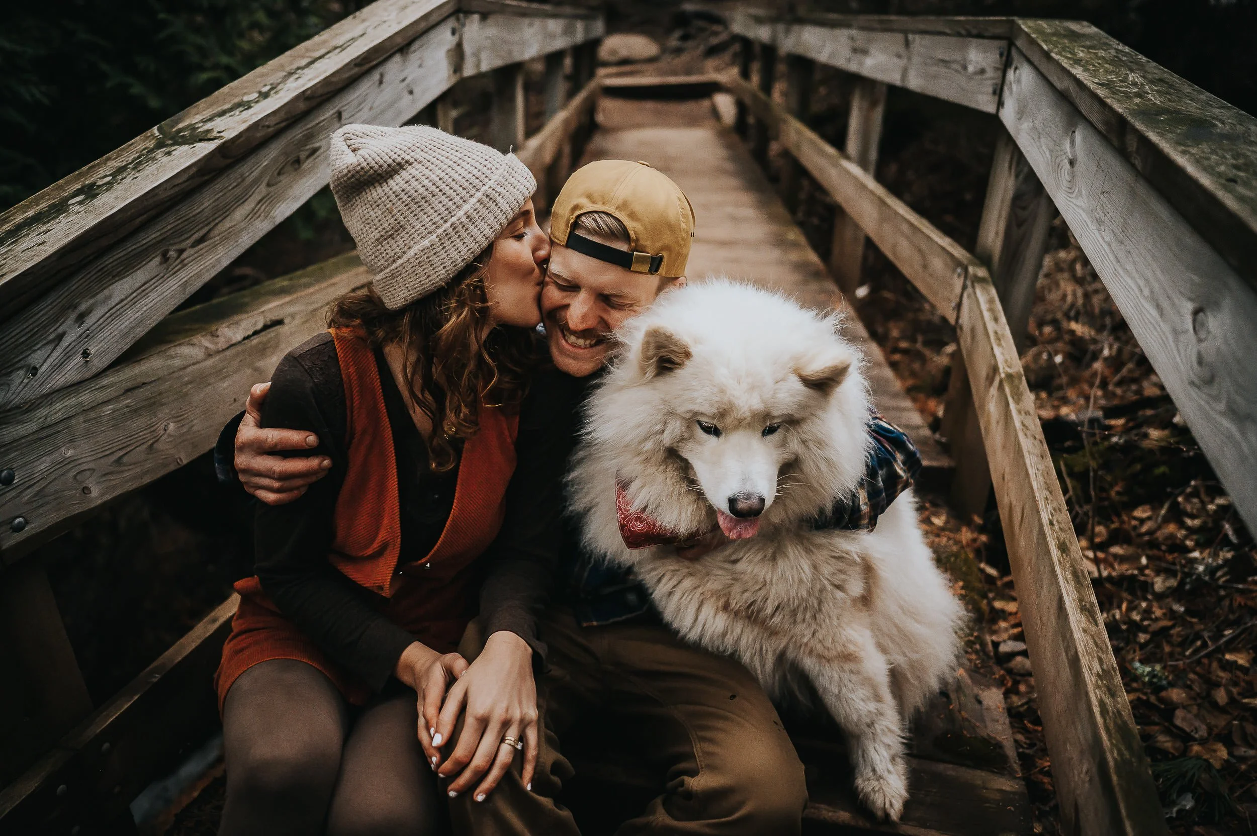A couple snuggles together with a large white fluffy dog in a rustic wooden structure, cozy and joyful.