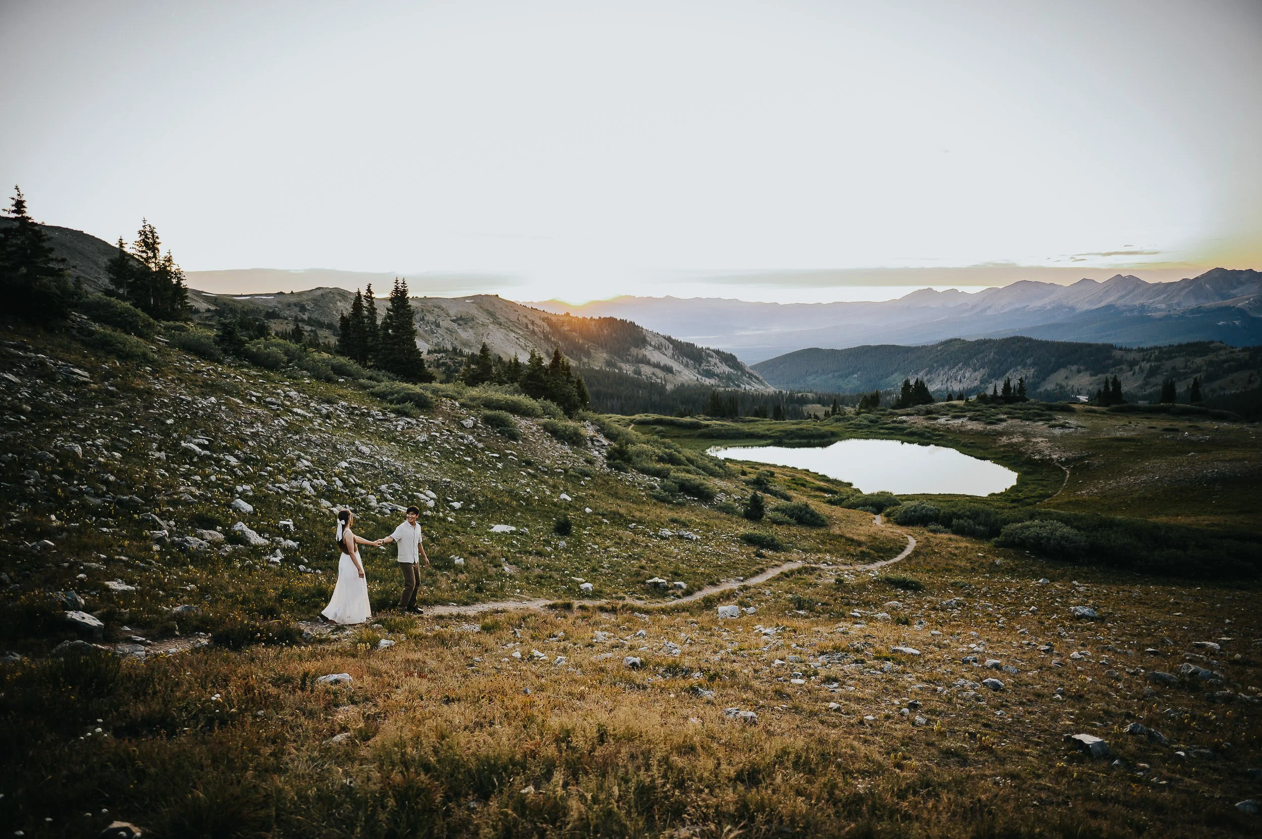 A bride and groom stand as tiny figures in a sweeping Colorado alpine meadow, mountains and a small lake behind them.