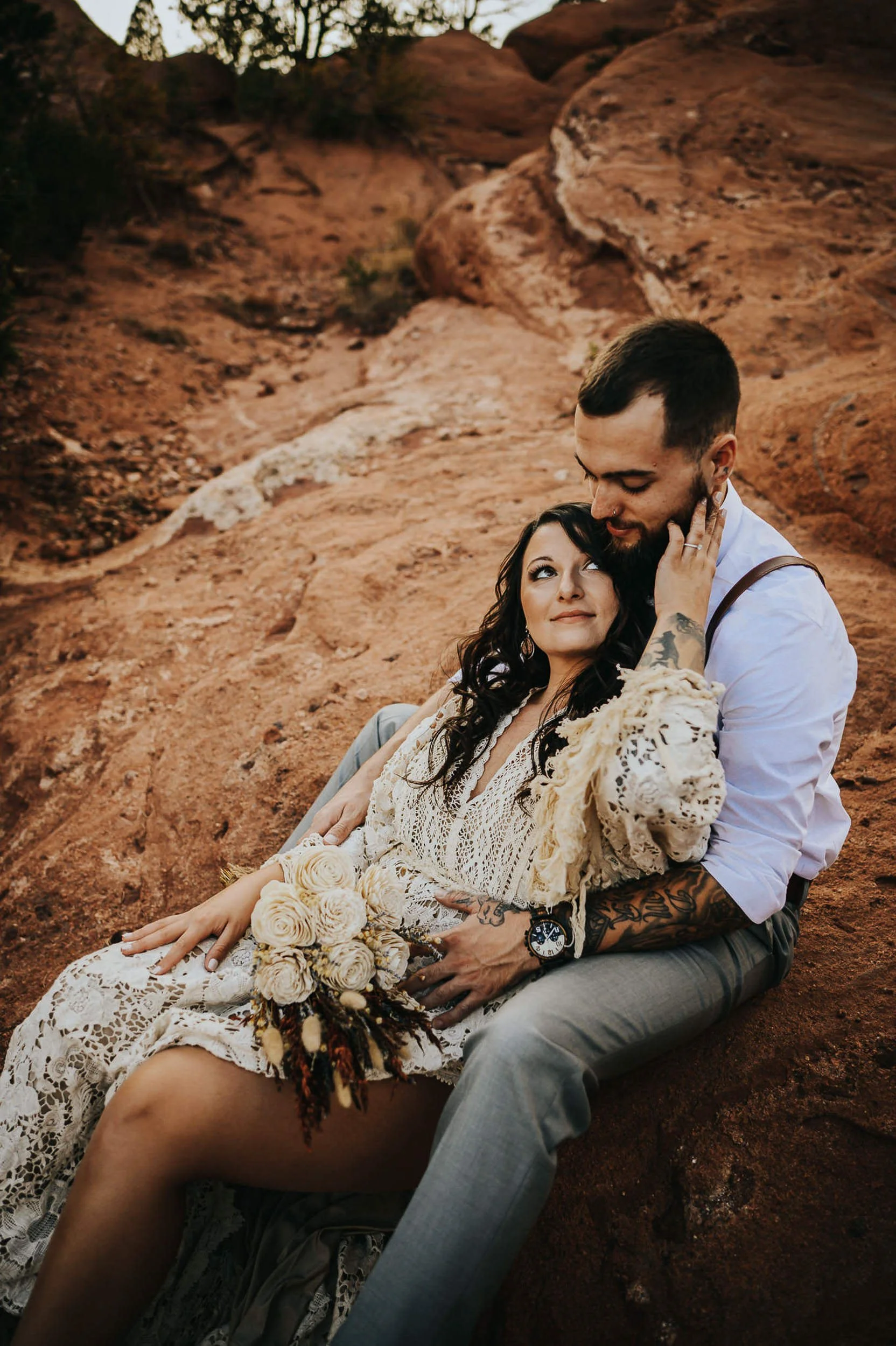 A couple sits close together on red rocks, wrapped in a cream blanket, faces tender and close.
