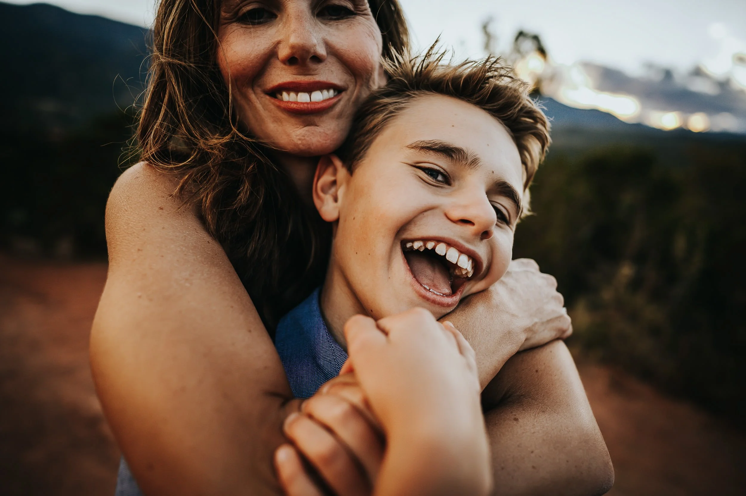 A mother and her son press cheek to cheek, both laughing with mountains behind them.