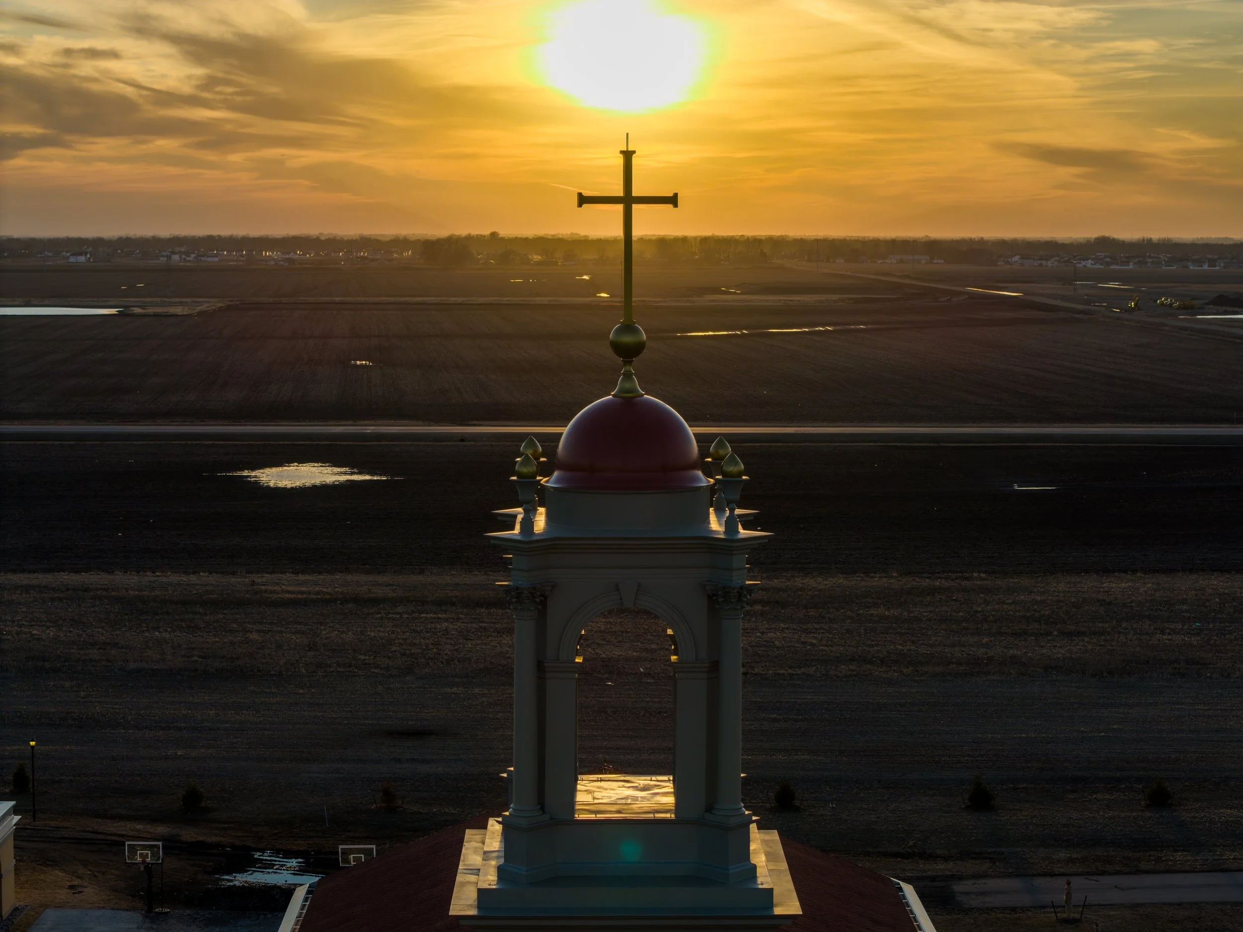 A church steeple with a cross at the top, silhouetted against a sunset or sunrise over flat farmland.