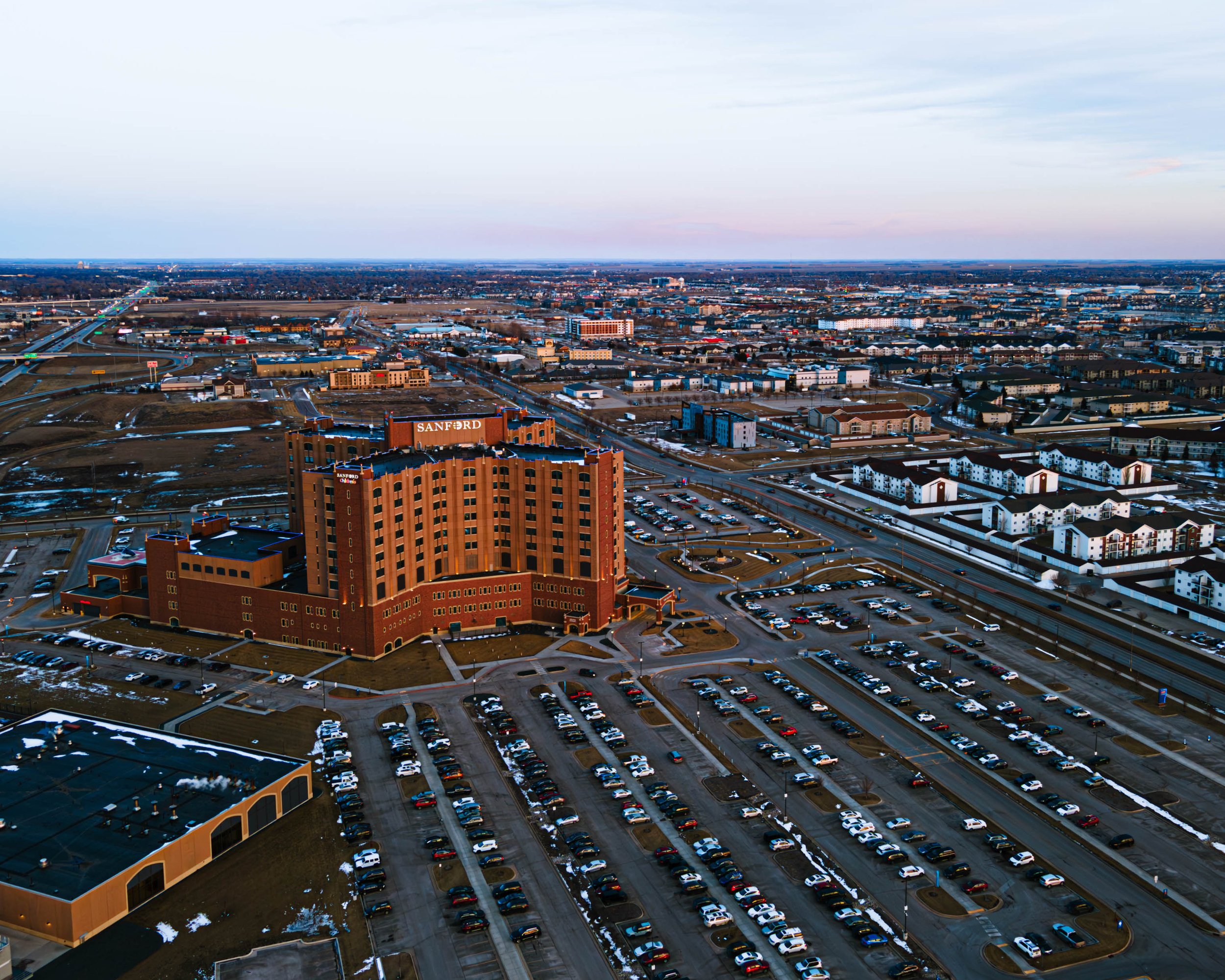 An aerial view of a cityscape at dusk showing a large brick hospital building labeled 'SANFORD' surrounded by parking lots, roads, and numerous residential and commercial buildings extending into the distance.