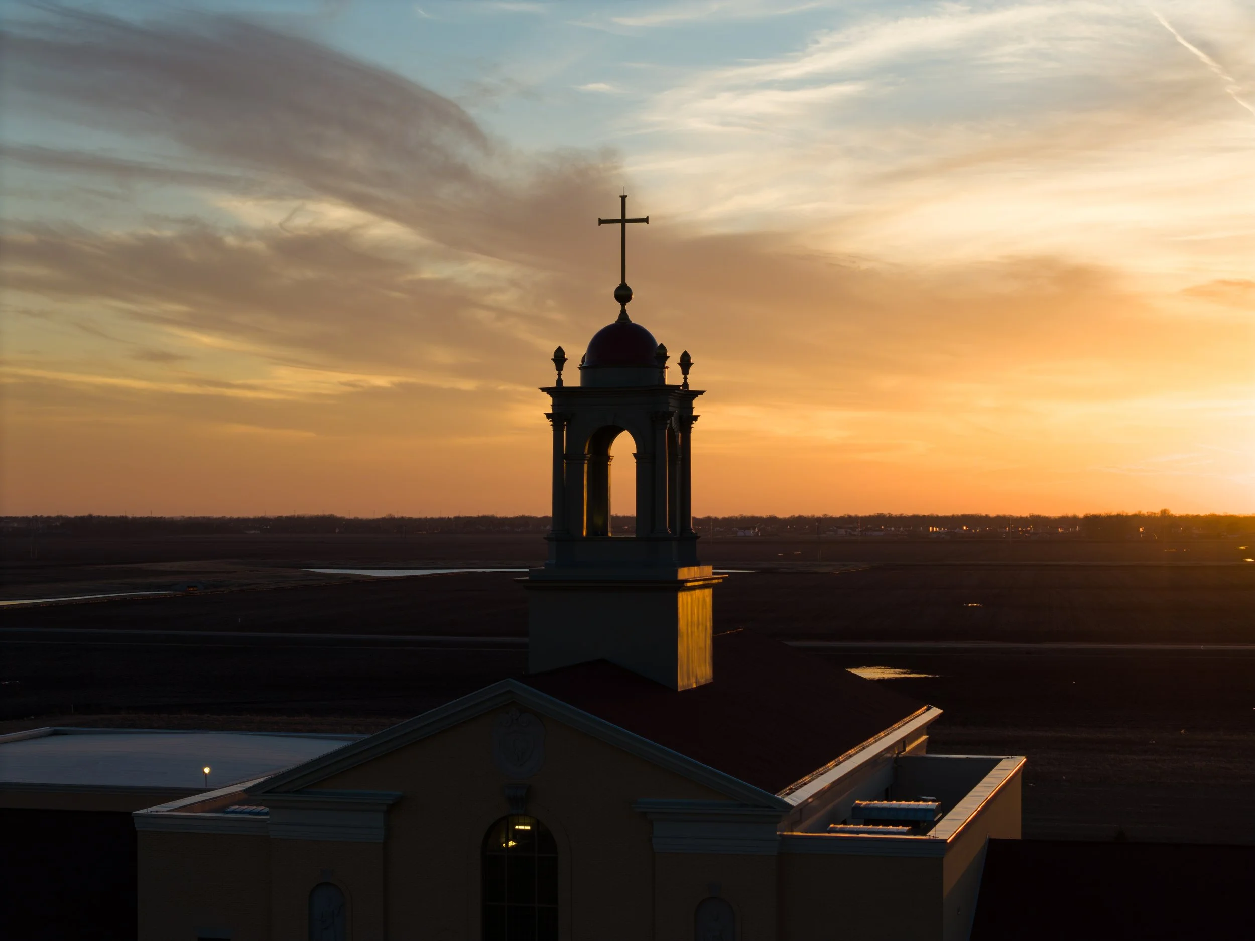A church steeple with a cross on top is silhouetted against a sunset sky with wispy clouds.