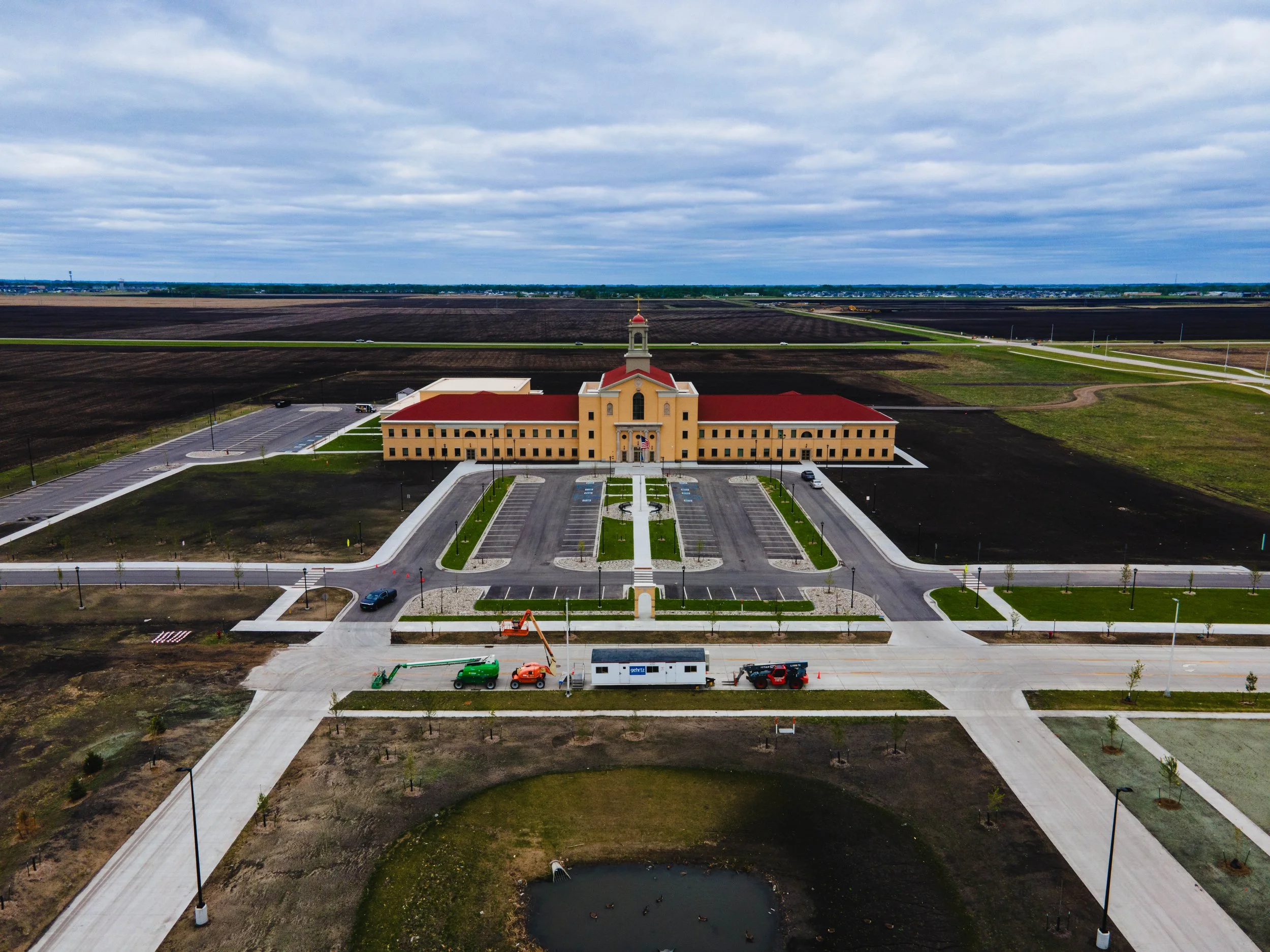 A large school building with a red roof and beige walls in an expanding development area, surrounded by parking lots, roads, and open fields with some construction activity visible.
