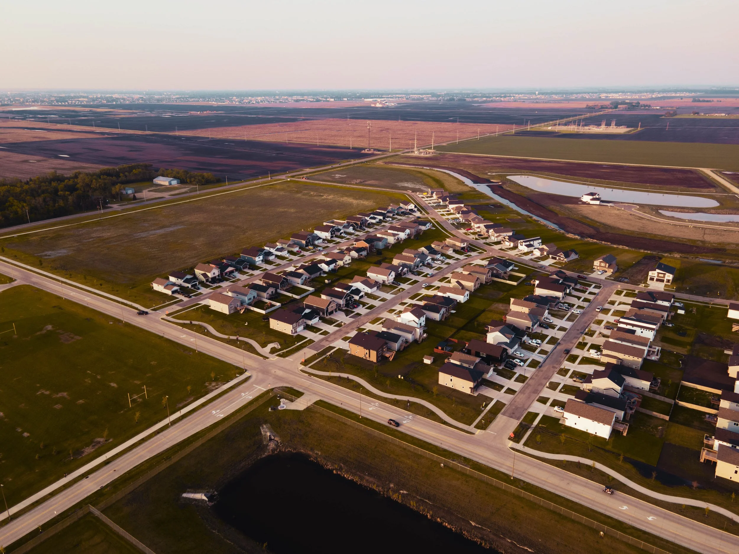 Aerial view of a residential neighborhood with houses and roads, near open fields, ponds, and farmland, during sunset.