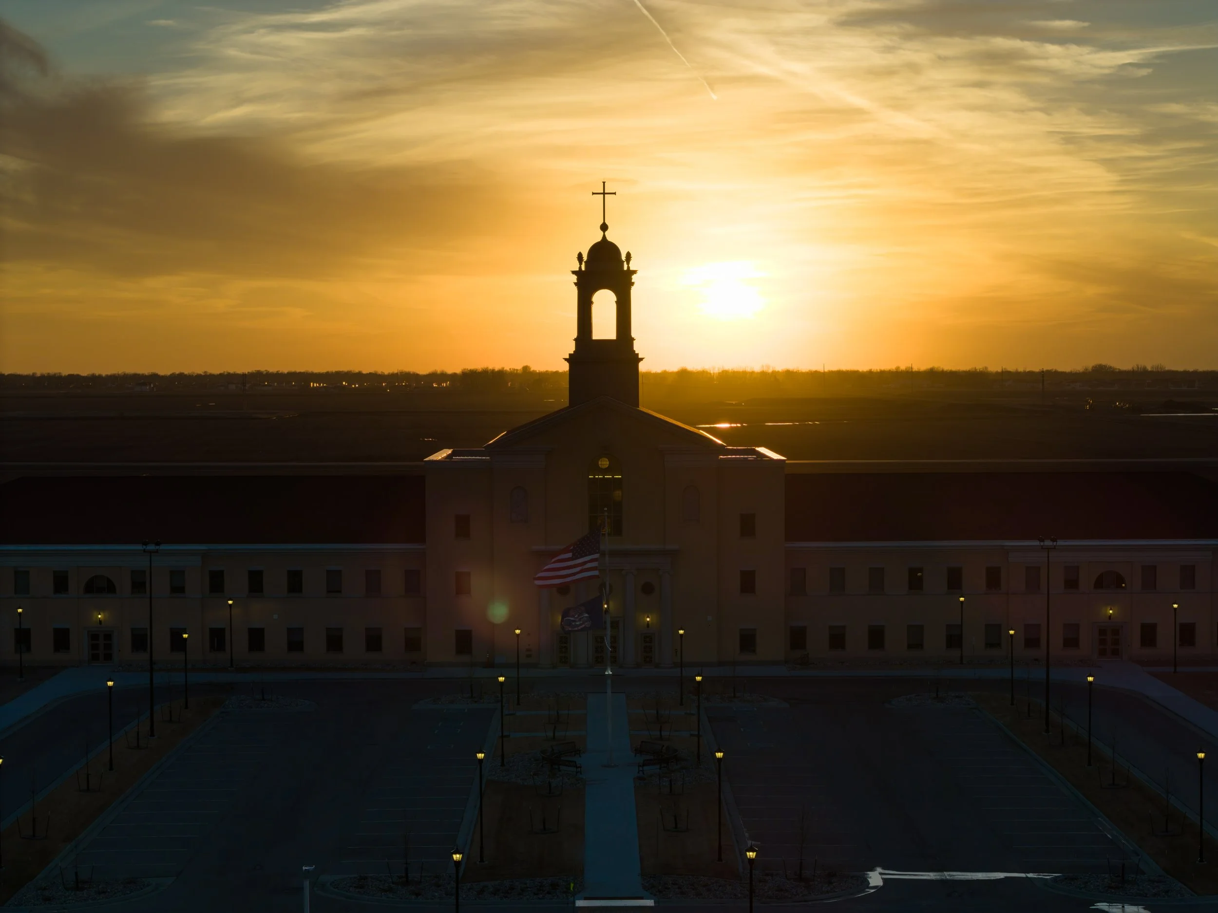 Sunset over a church with a steeple topped by a cross, American flags in front, and a parking lot with street lamps
