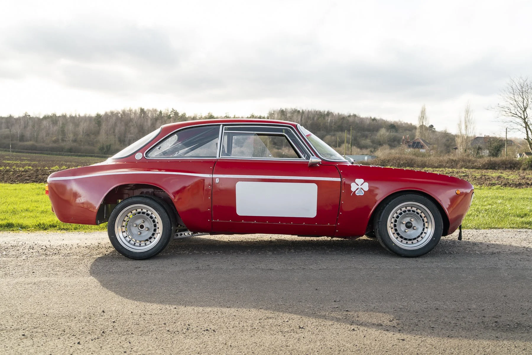 A vintage red race car with a blank white rectangle on the door, parked on a rural road with grassy fields and a cloudy sky in the background.