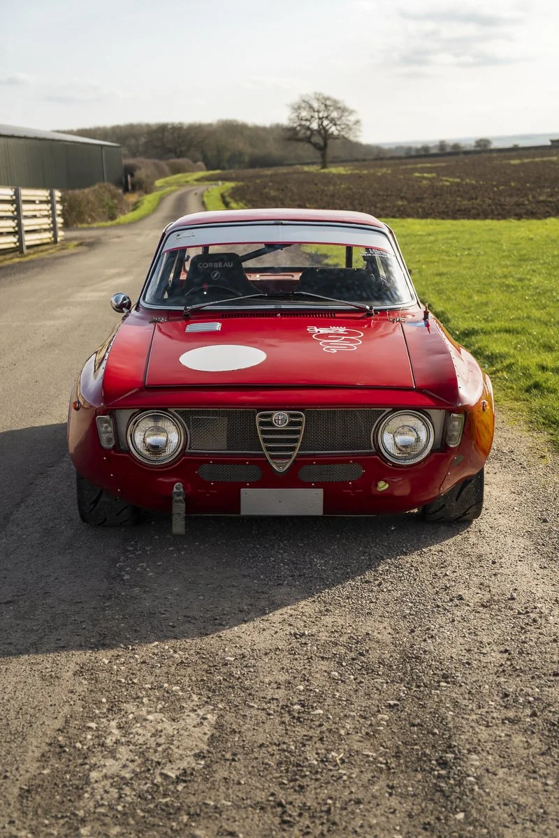A vintage red Alfa Romeo car parked on a country road with fields and trees in the background.