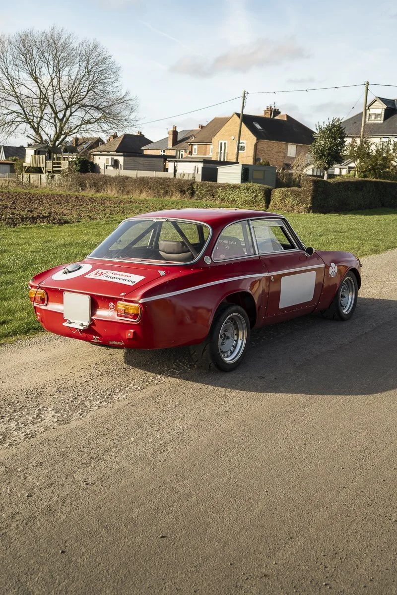 A vintage red race car parked on a dirt road in a residential neighborhood with houses and trees in the background.