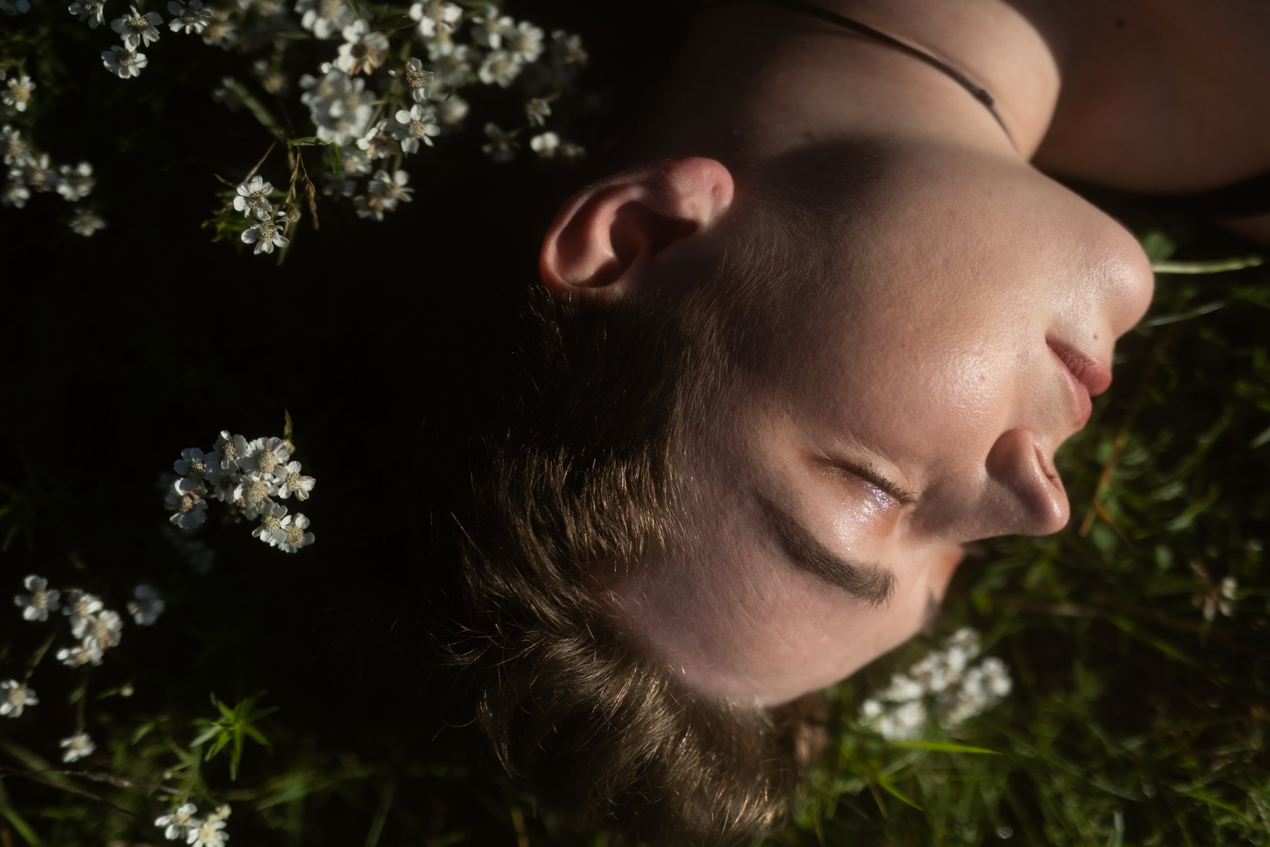 Close-up of a person lying down outdoors with their eyes closed, surrounded by small white flowers and greenery.