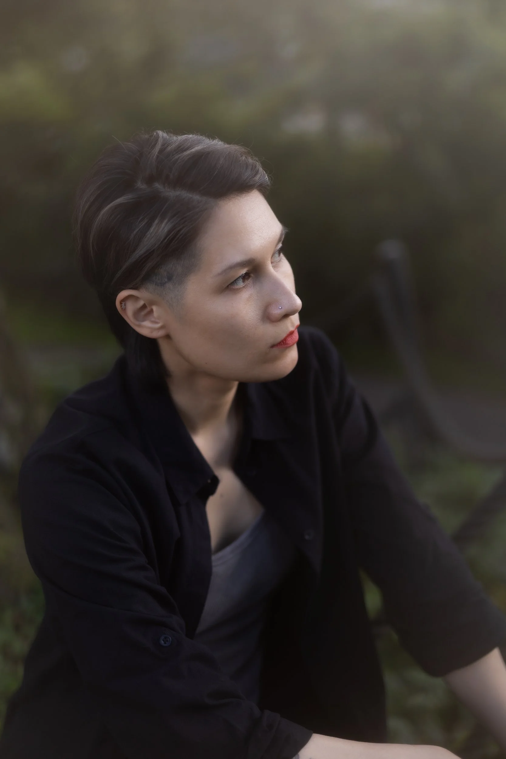 A person with short, dark hair styled with an undercut, wearing a black jacket, sitting outdoors with blurred trees in the background, looking to the side.