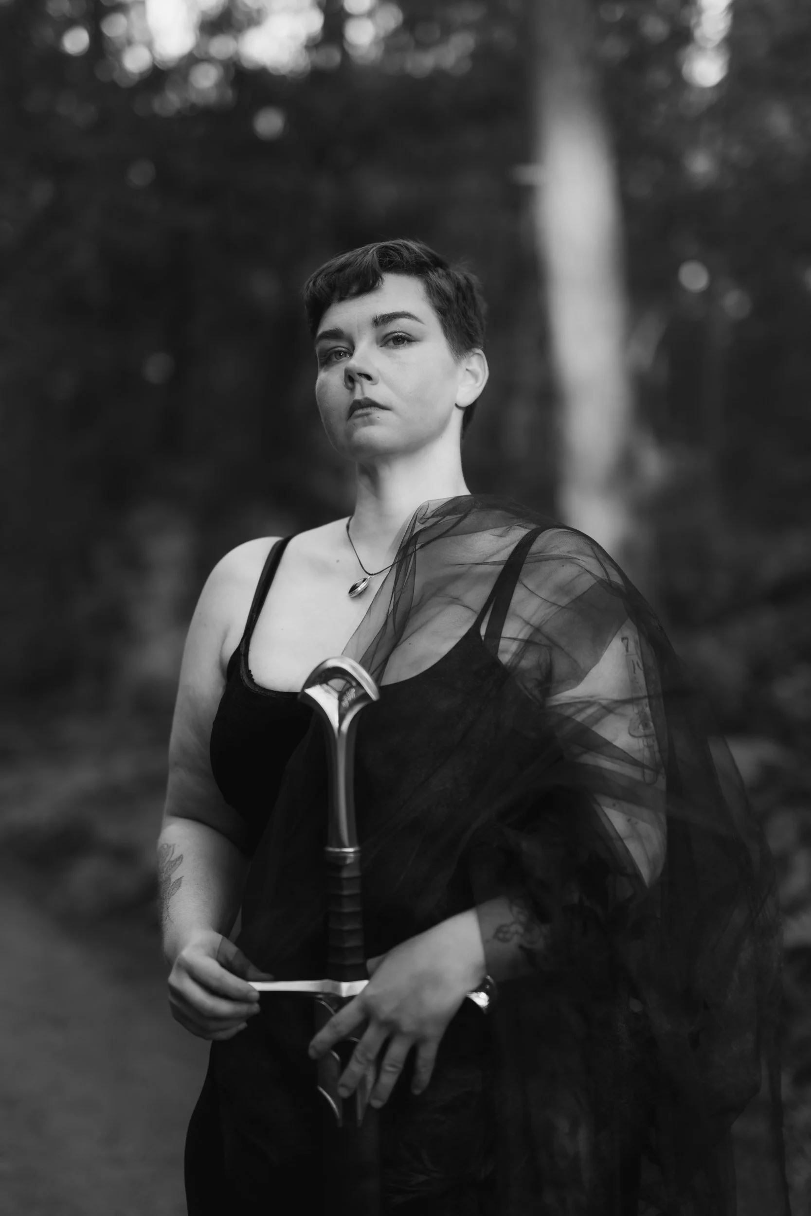 A black and white portrait of a person with short dark hair, wearing a black top with thin straps, a sheer black shawl, and a necklace. They are  outdoors with blurred trees in the background, holding a sword.