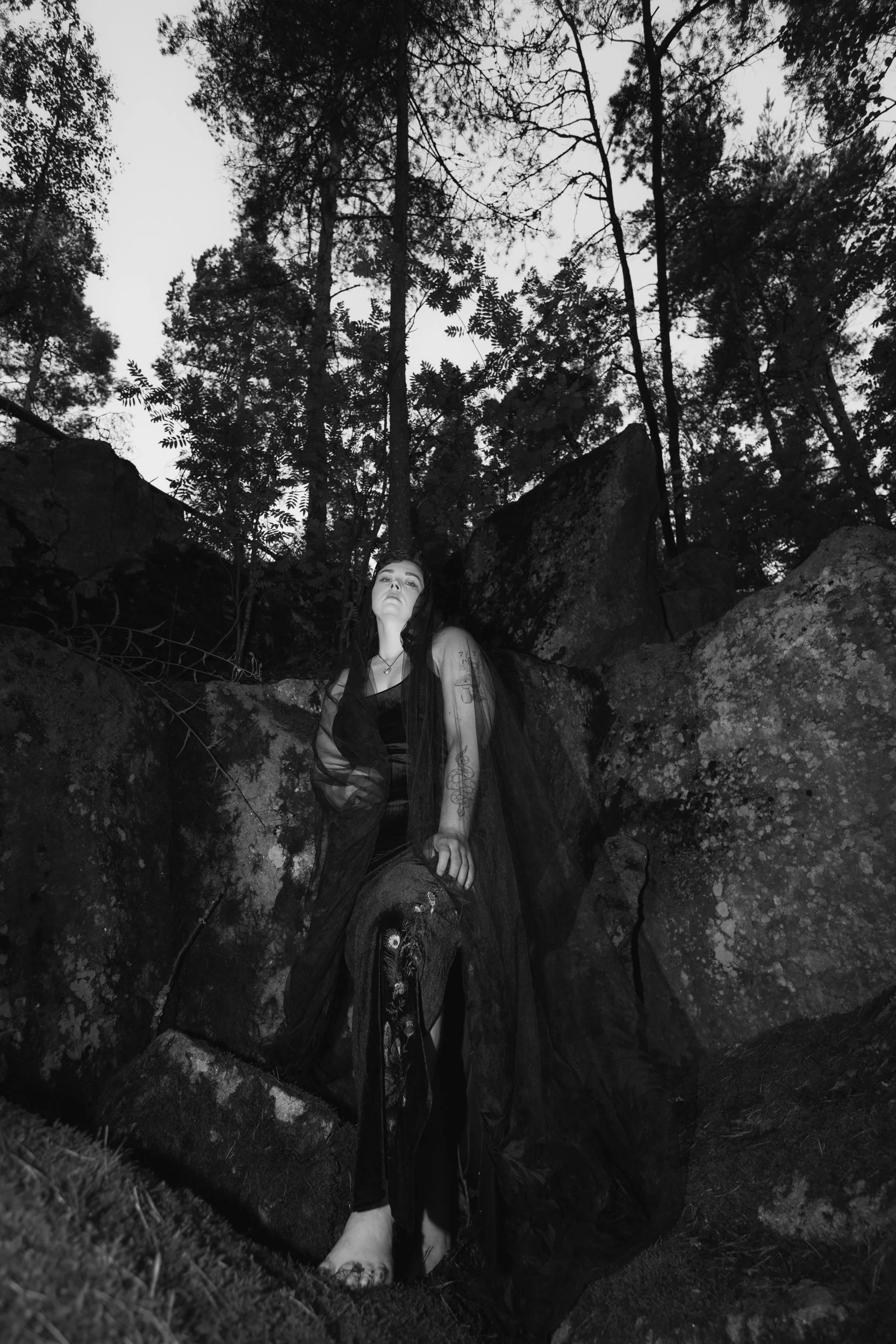 A person with long hair and tattoos sitting on rocks in a wooded outdoor area during the daytime, with trees visible in the background, captured in black and white.