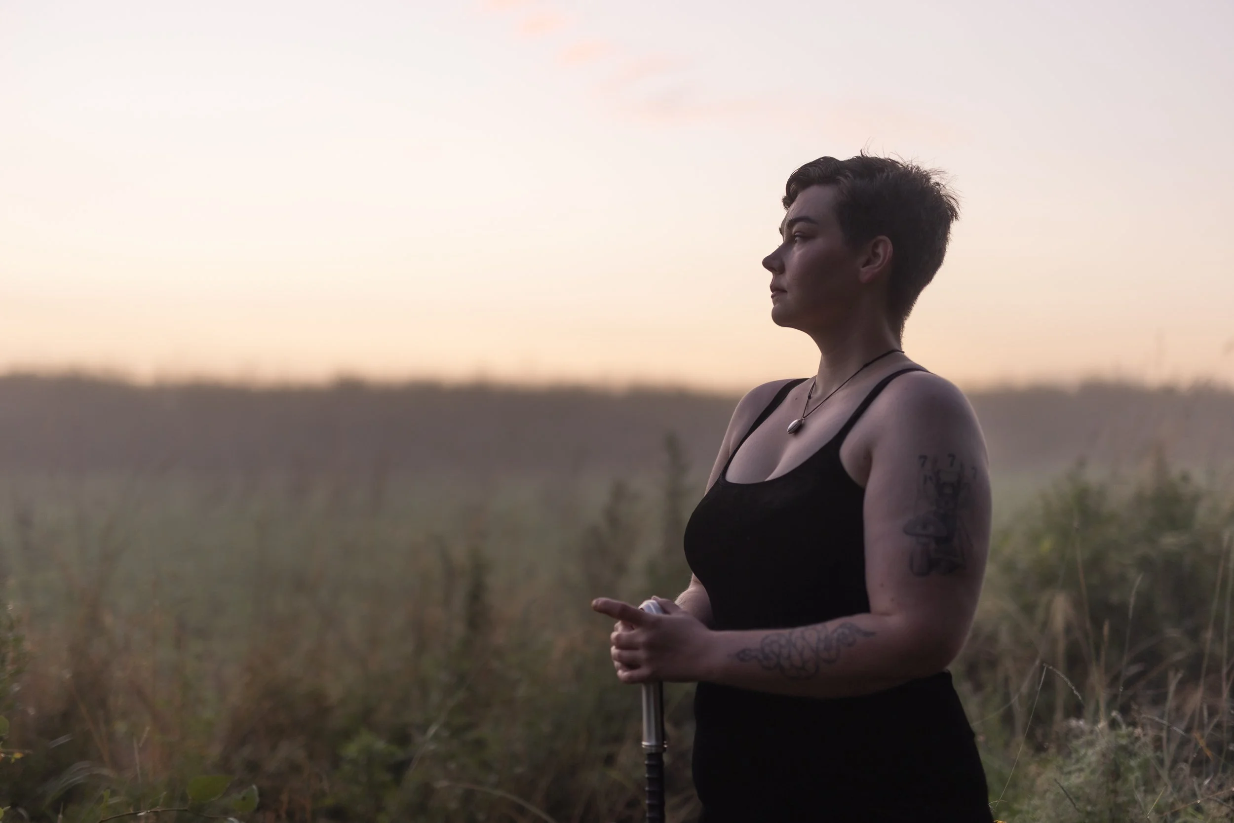 A person with short dark hair and tattoos on their arm stands outdoors in a field during sunset, looking to the right, wearing a black tank top and a necklace.