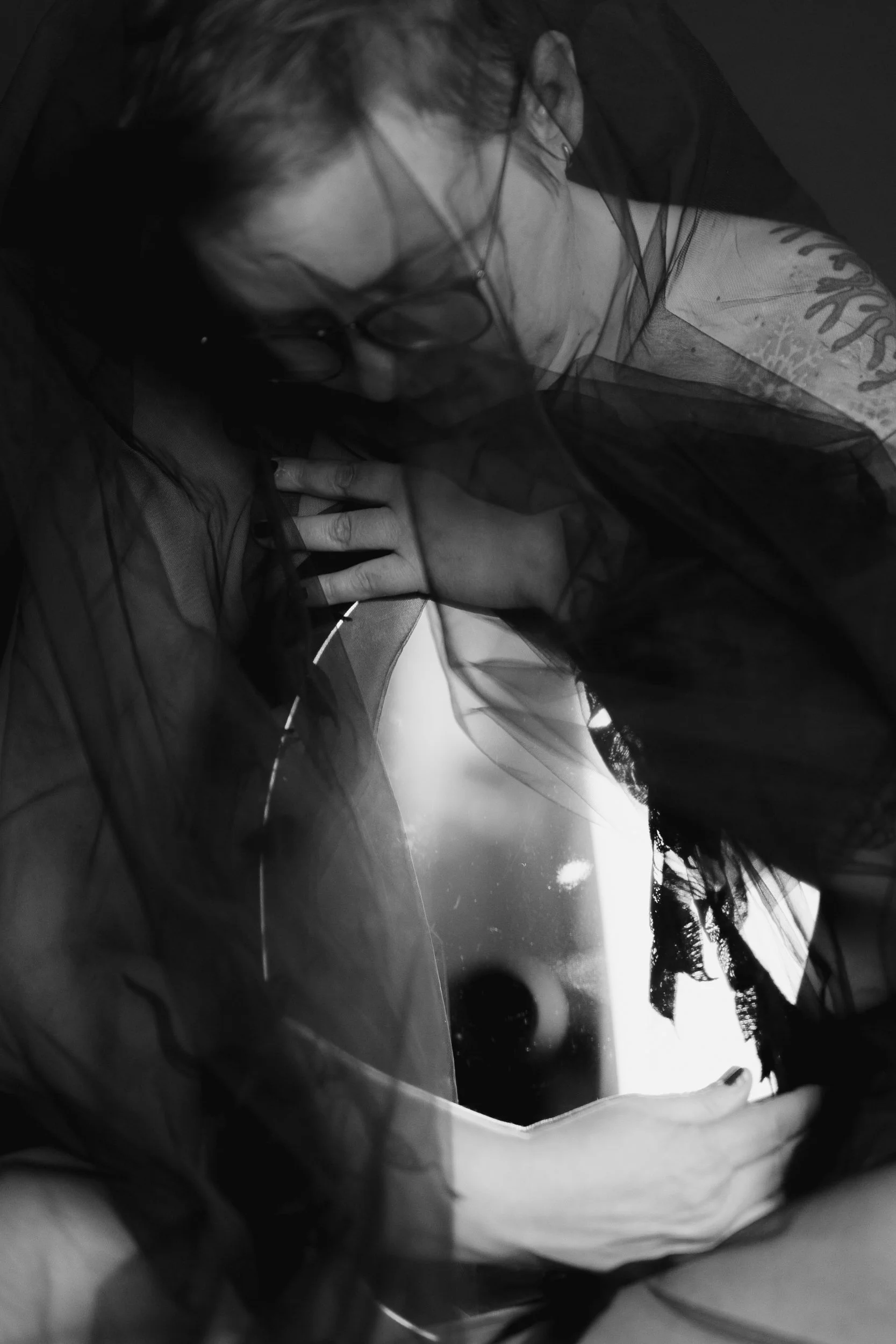 Tinksu is sitting under a black veil and holding a kidney-shaped mirror on their lap. They hug the mirror. The vibe is sad, amotional and also calm