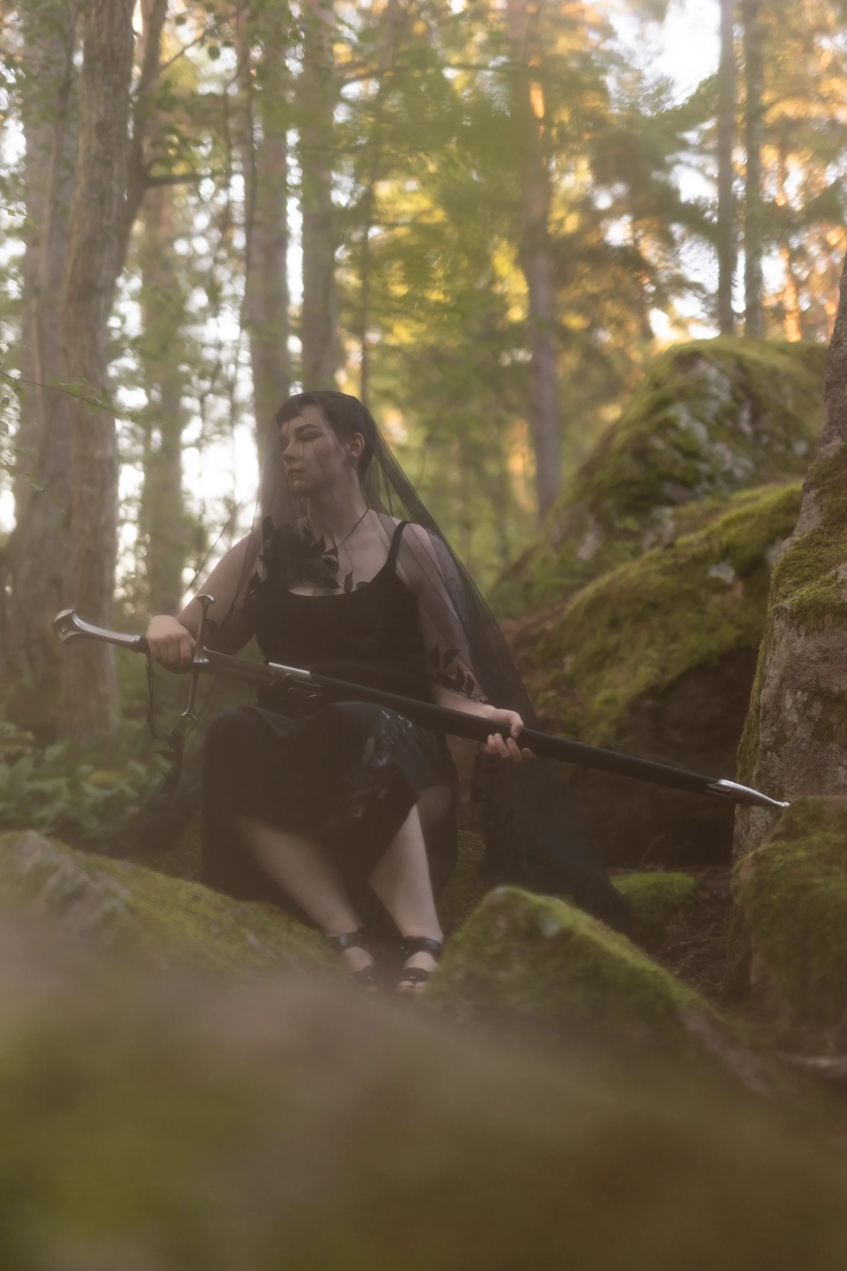 A person in black clothing and a veil, holding a sword, sitting in a mossy forest with tall trees and warm sunlight filtering through the leaves.