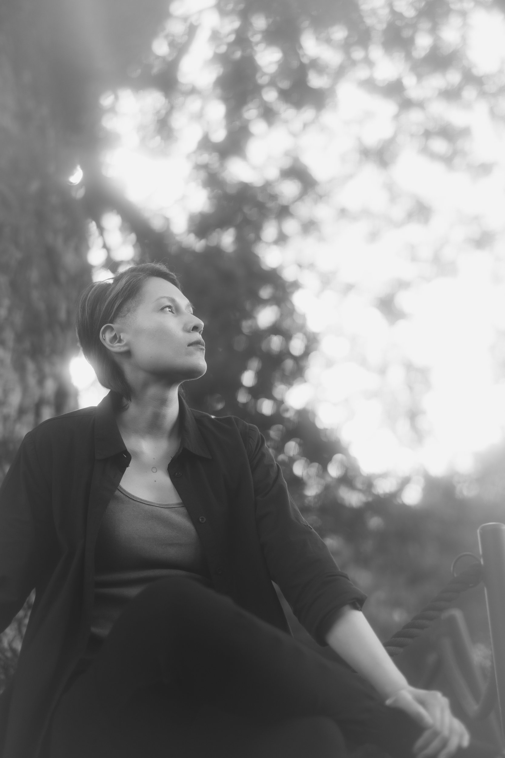 A person with short hair, wearing a dark jacket and a light-colored top, is sitting outdoors in nature. They are looking to their left with a thoughtful expression. The background shows trees and dappled sunlight, creating a peaceful atmosphere.