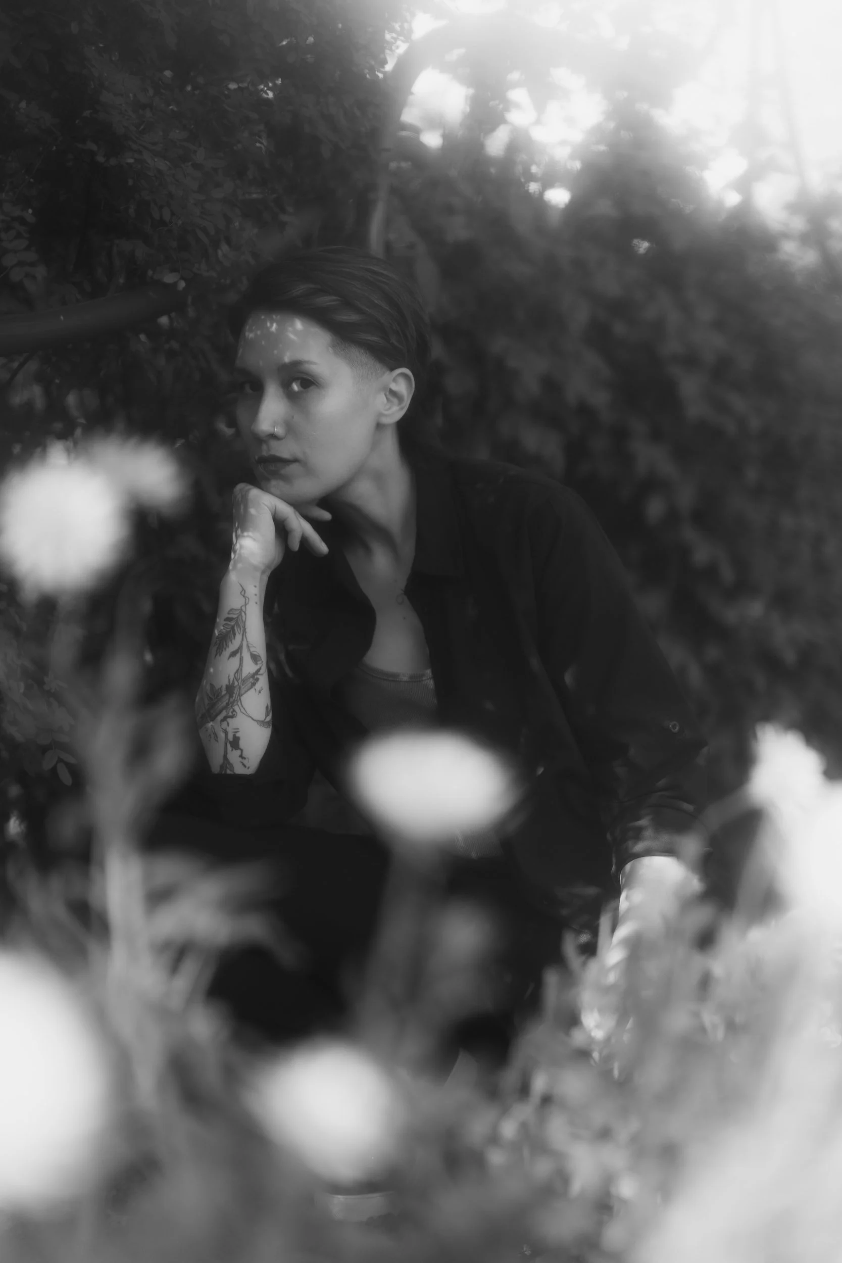 Black and white portrait of a person with short hair, tattoos on their arm, and a nose piercing, sitting outdoors among blurred plants.