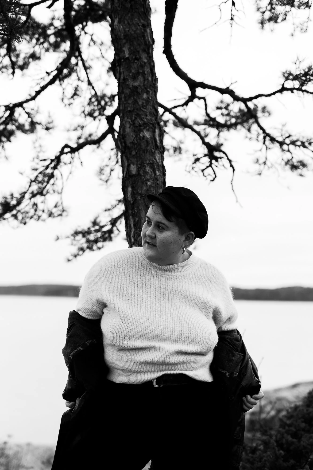 Siiri is leaning on a tree trunk and looking right. The sea is behind them. They are wearing a white woollen shirt and a cap.