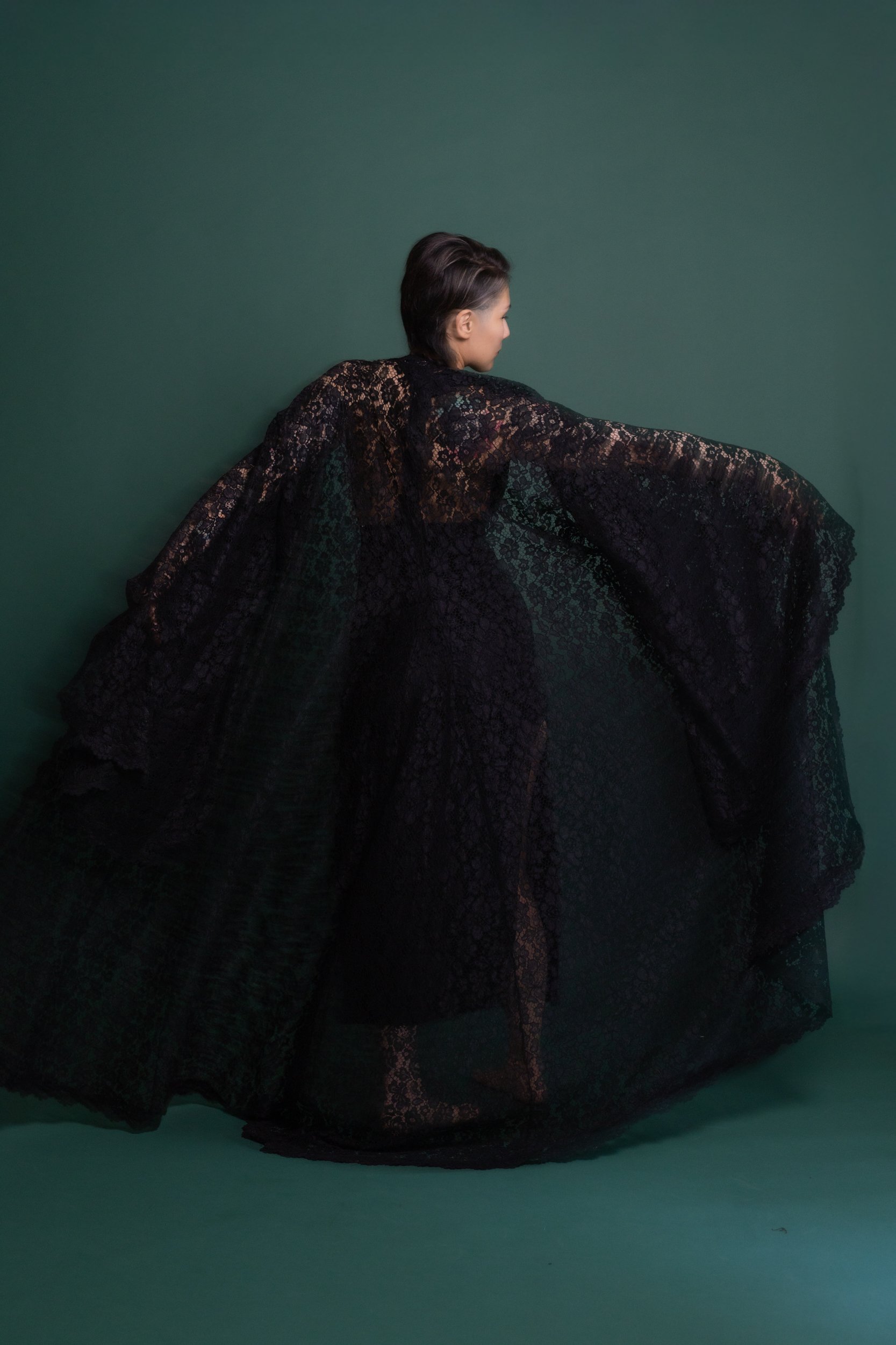 A person with dark hair in a slicked-back style wearing a flowing black lace dress interwoven with floral patterns.