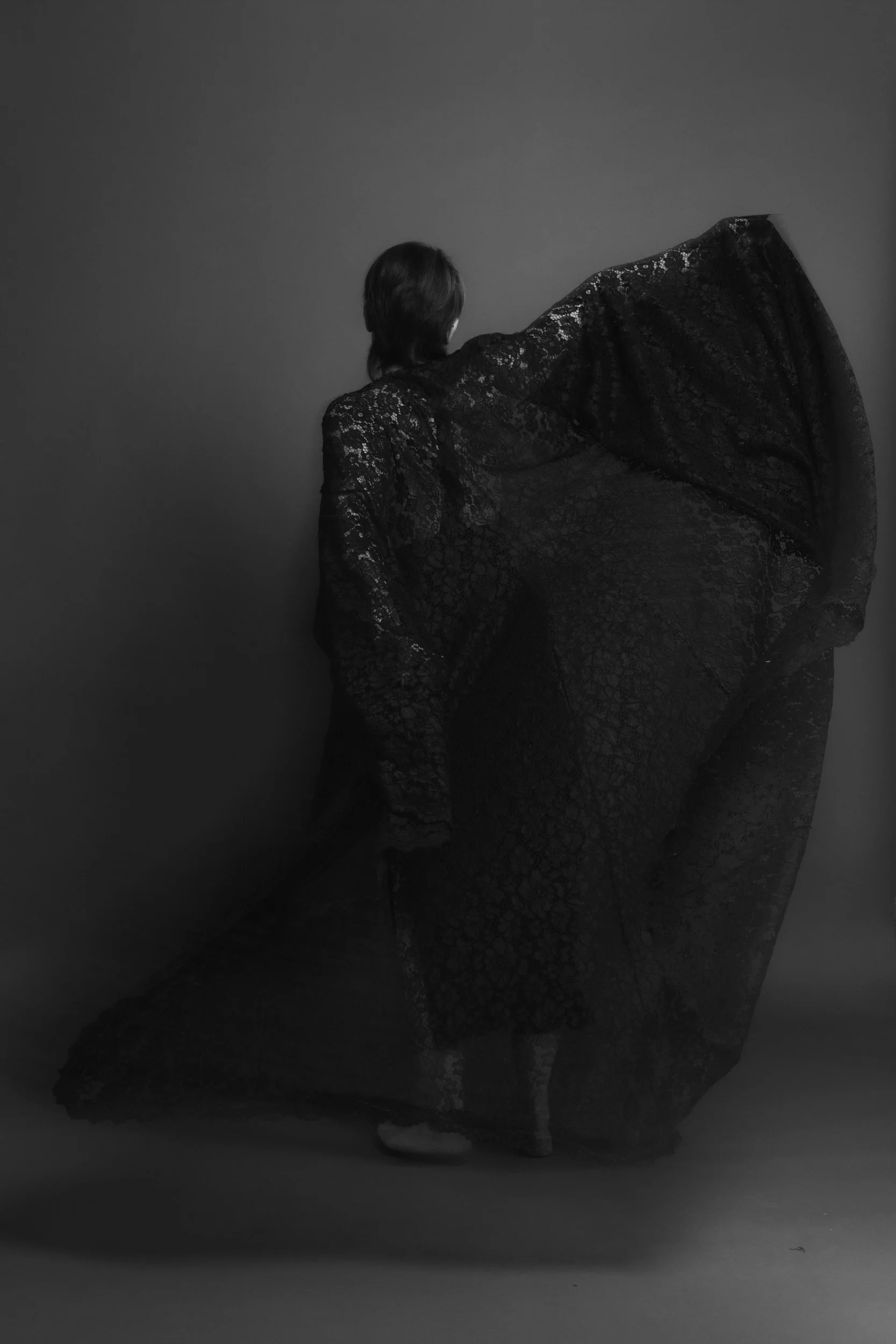 A person with short hair in a lace dress, facing away, standing in front of a plain wall.