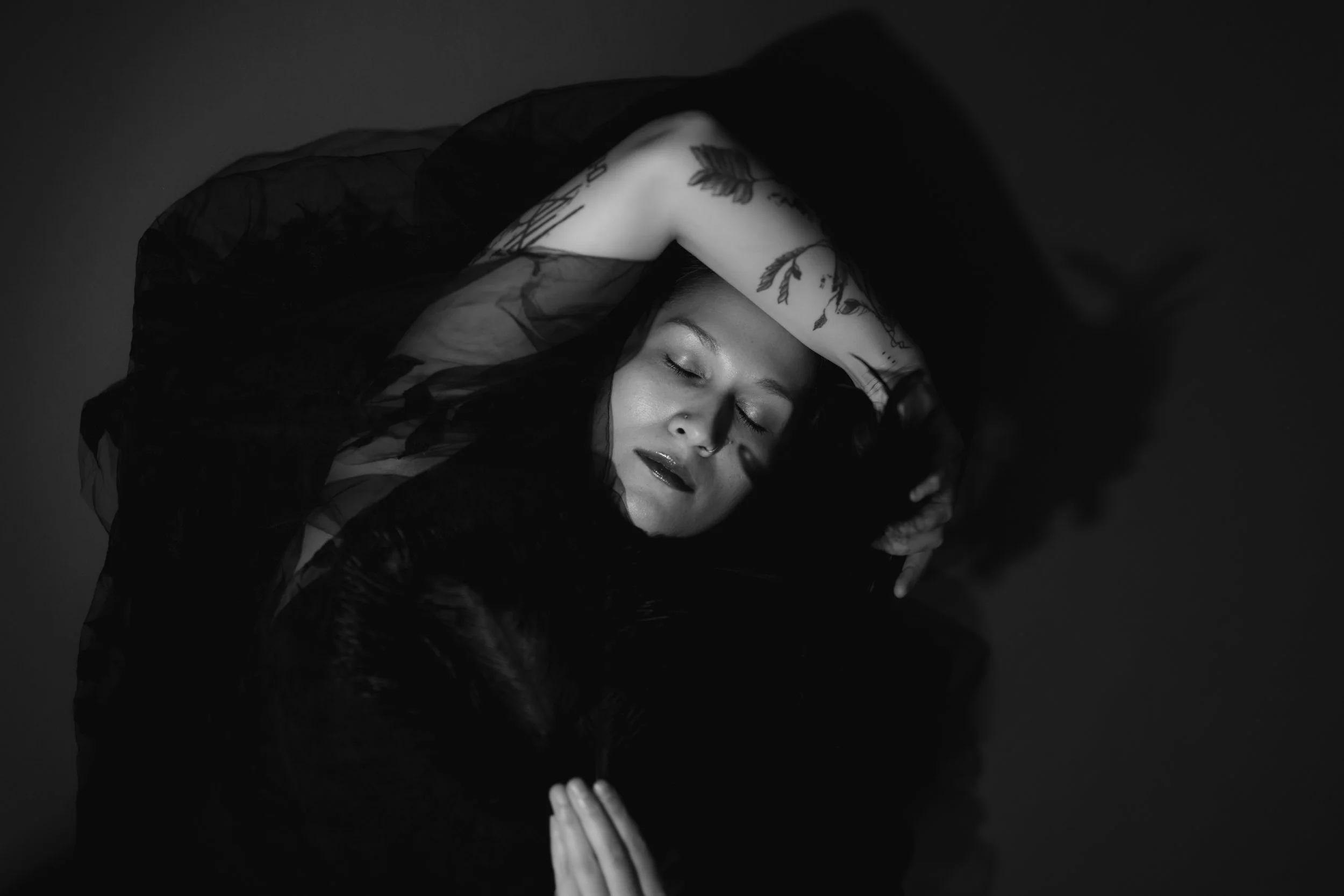 A black and white photo of a person with closed eyes, resting their head on their folded arm, with tattoos visible on their arm and shoulder, and a sheer black fabric draped over them.