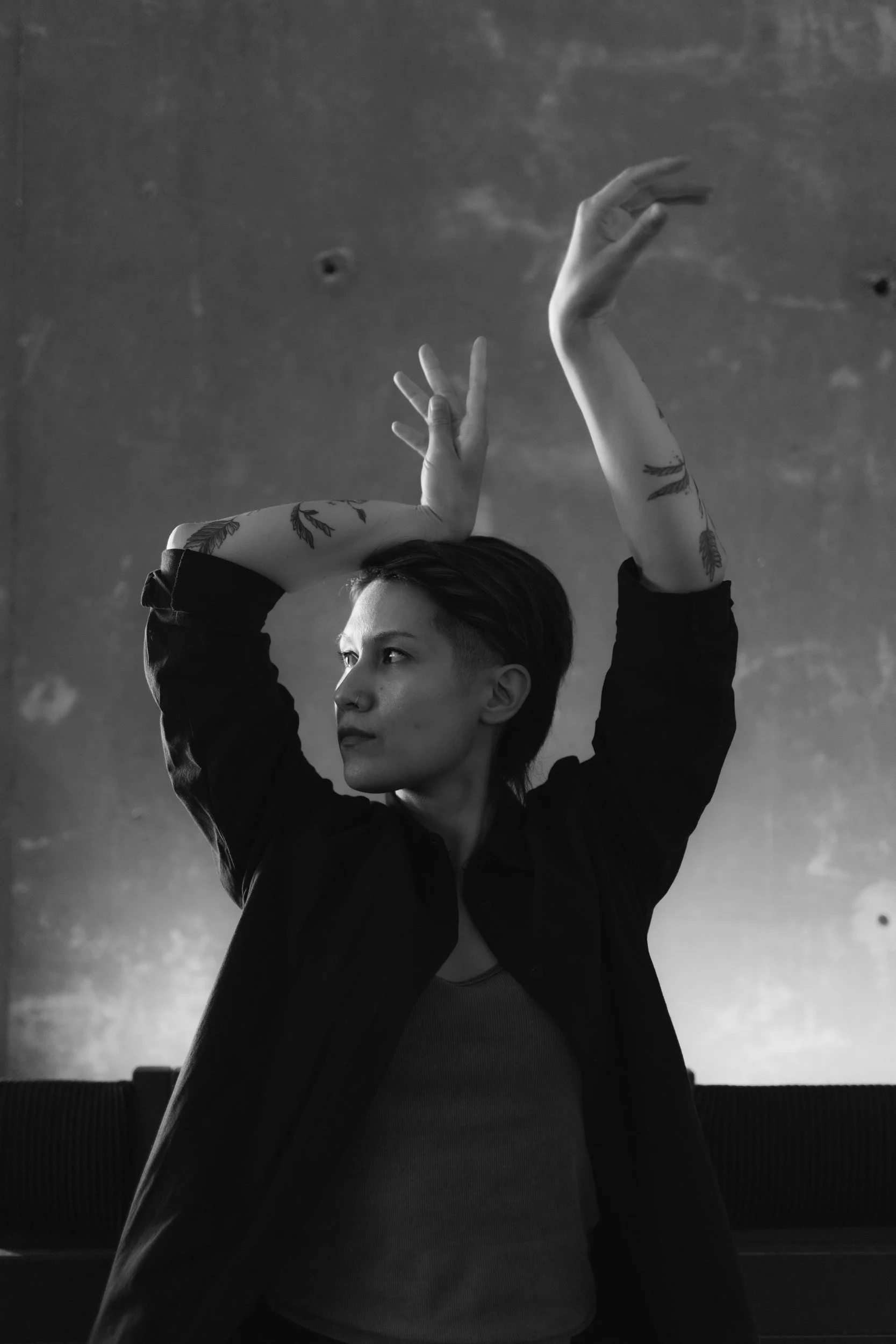 Black and white photo of a person with short hair and tattoos on both arms, raising her left hand above her head, with a serious expression, in front of a textured wall.