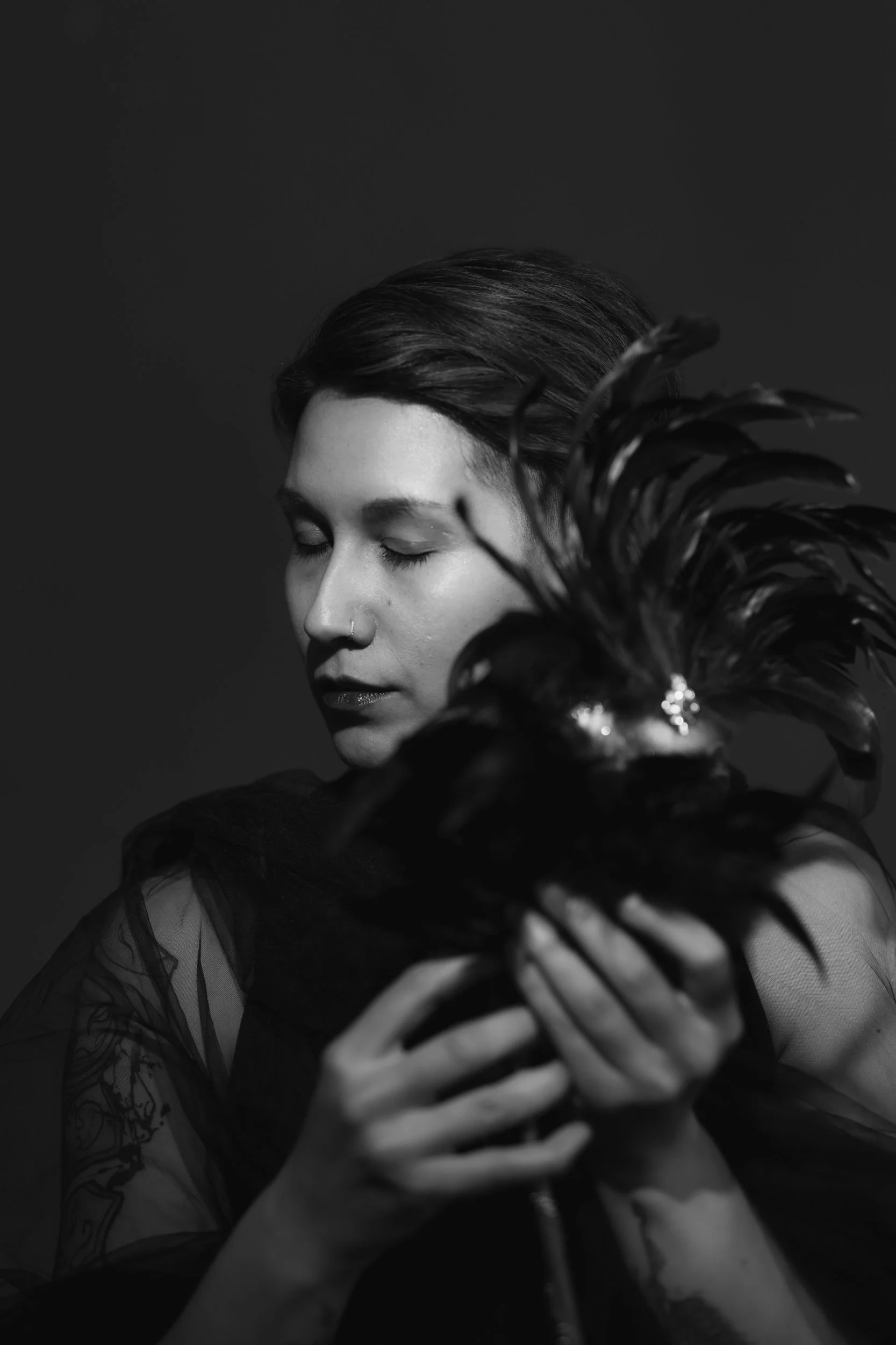 A black and white photograph of a person with closed eyes and short dark hair, holding a dark feathered plant close to their face. The person has a nose piercing and tattoos visible on their arms.