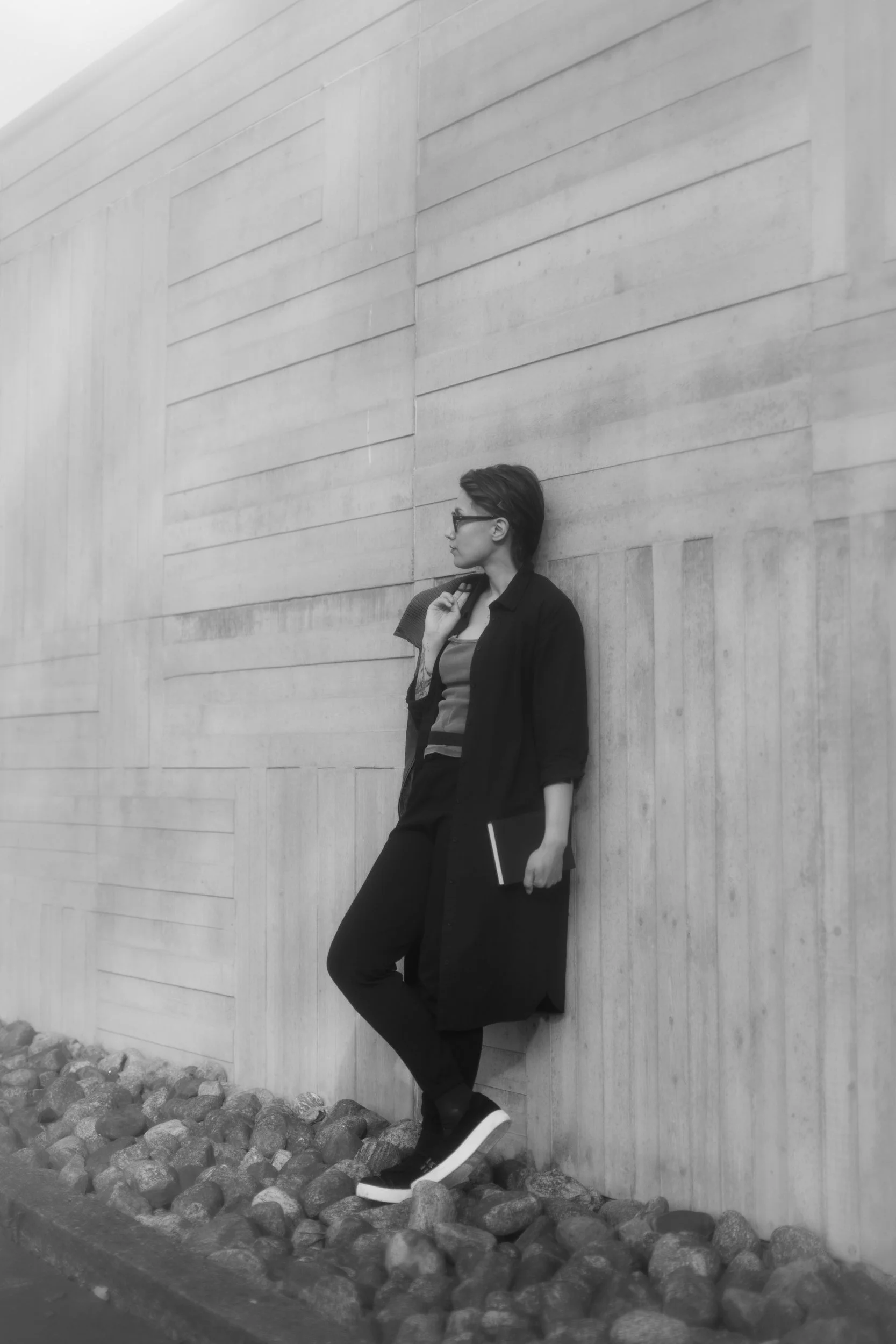 A person with short hair and glasses leaning against a large concrete wall, holding a notebook and a pen, standing on rocky ground.