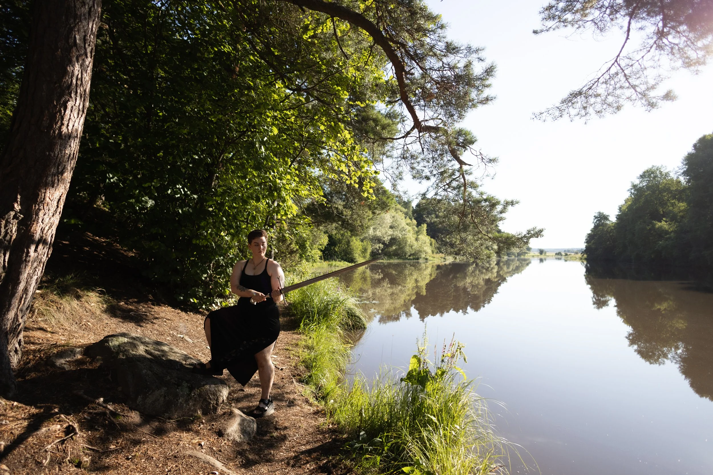 A person in a black dress sits by a riverbank under a large tree, holding a fishing rod with the river and trees reflecting in the water on a sunny day.