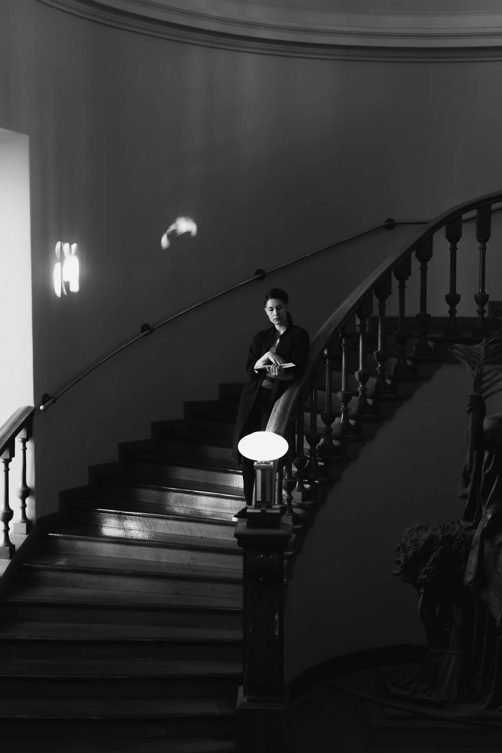 A black and white photo of a person leaning on a staircase railing, reading a book. The staircase has a curved wooden handrail, and there are decorative wall sconces providing light.
