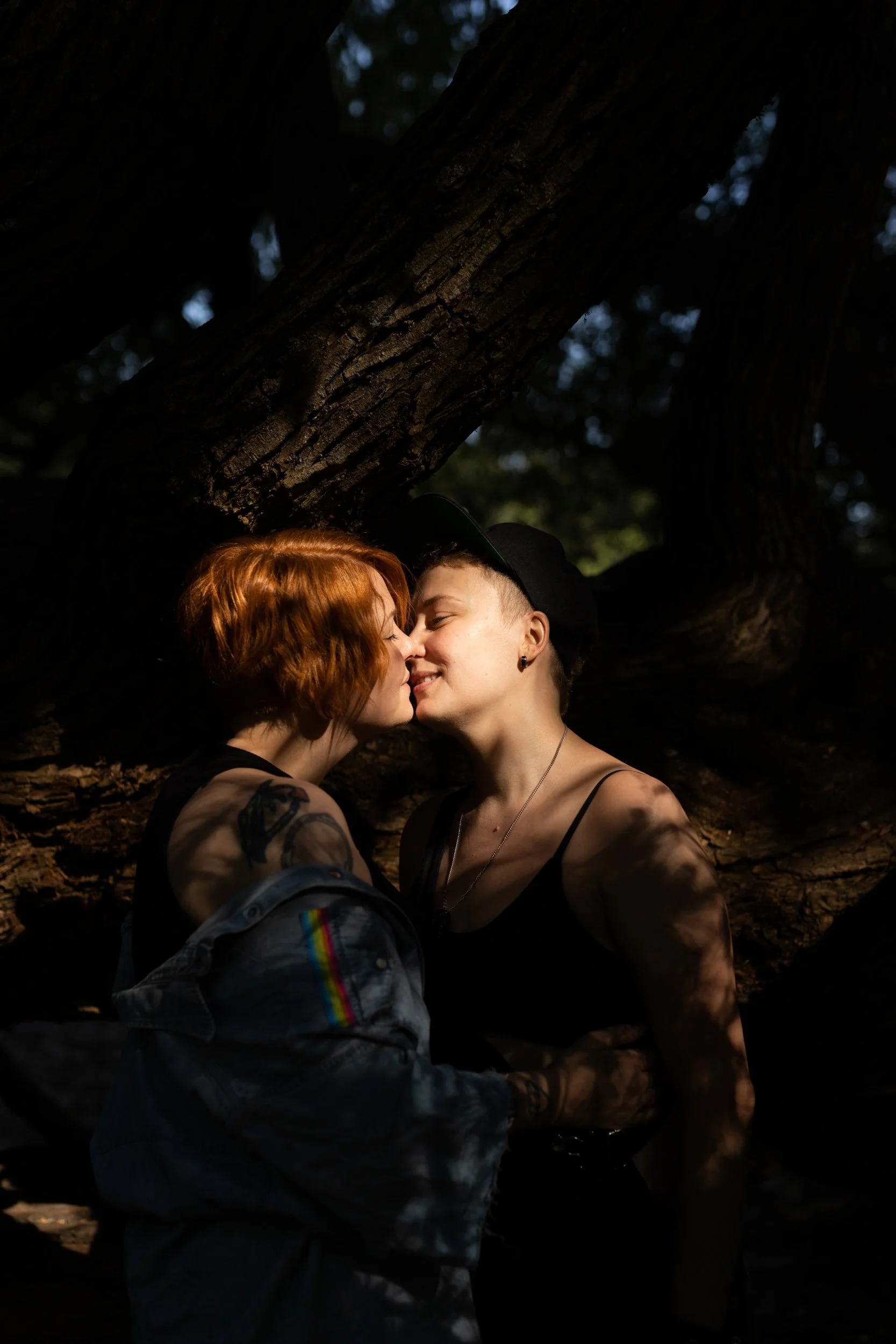 A lesbian couple kissing. They stand in front of a tree and beautiful golden hour light shines on them. They look very happy and very in love.